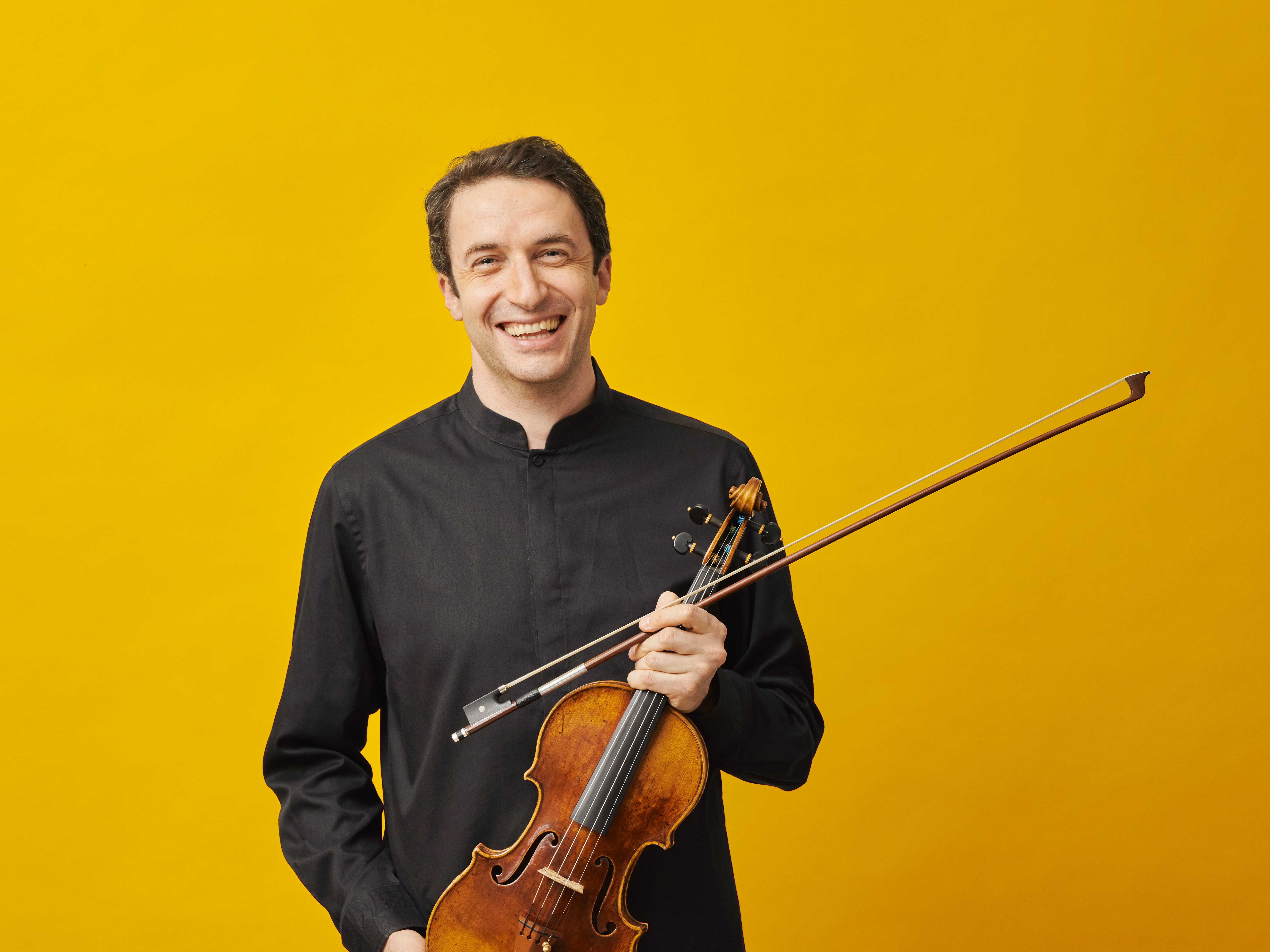 Konzertmeister Noah Bendix-Balgley lächelt herzlich in die Kamera und hält dabei seine Geige locker in der Hand. Er trägt ein schwarzes Hemd und hat hellbraune Haare.