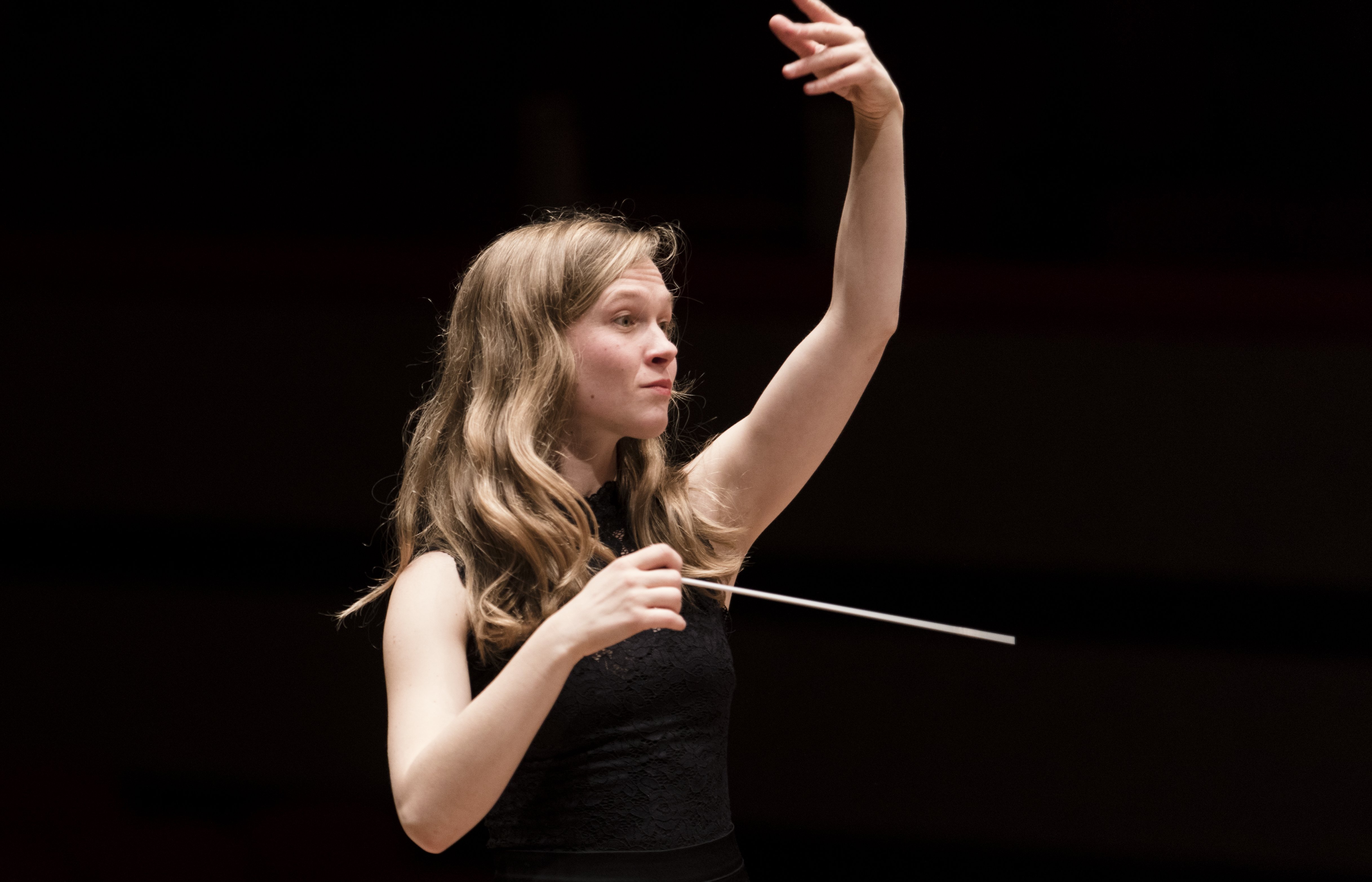 Eine Frau mit langen Haaren dirigiert ein Orchester, hält einen Taktstock in einer Hand und hebt die andere Hand. Sie trägt ein ärmelloses schwarzes Oberteil vor einem dunklen Hintergrund.