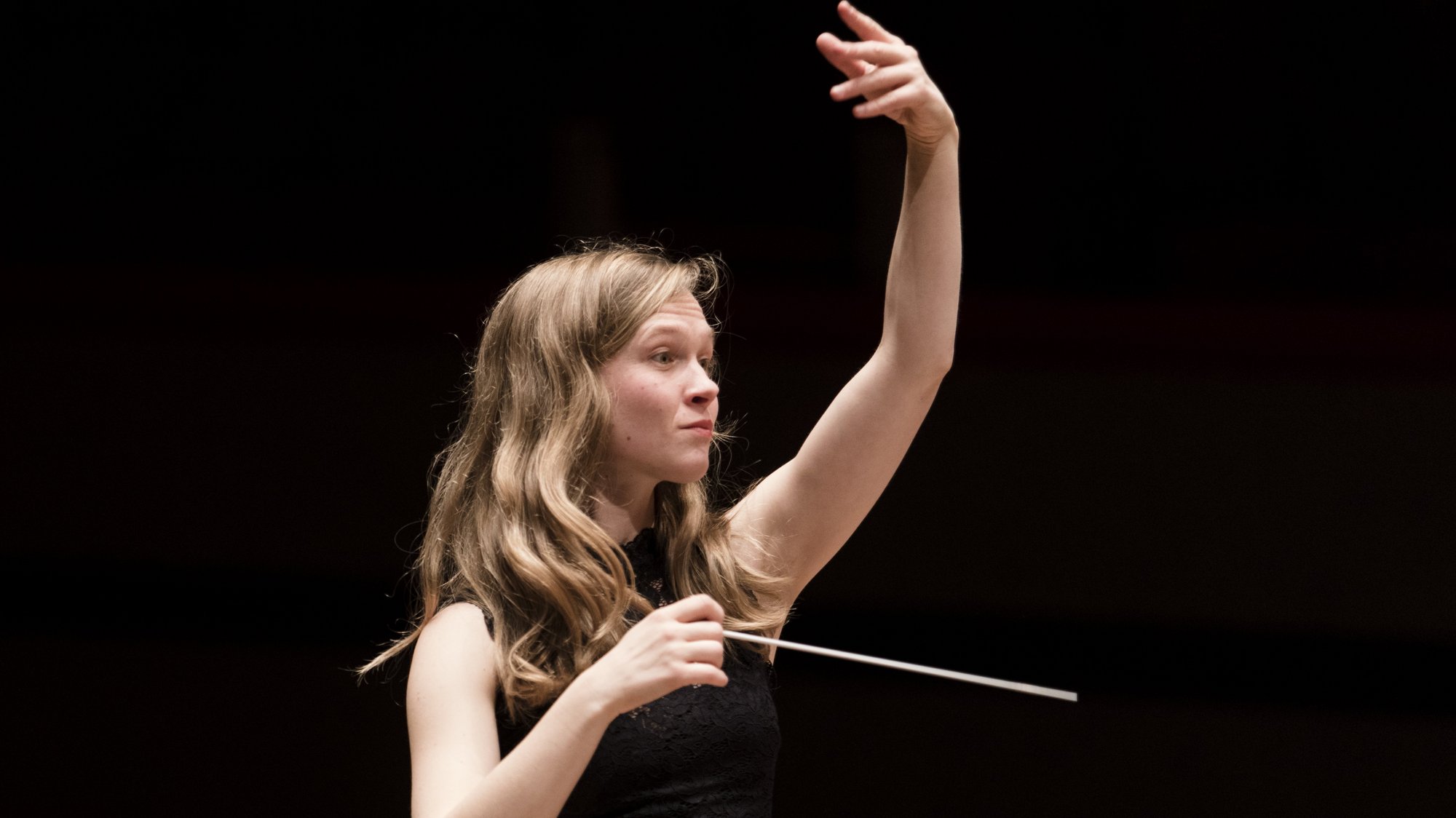 A woman with long, light brown hair conducts with a baton in her right hand, raising her left hand expressively against a dark background. She wears a sleeveless black top.