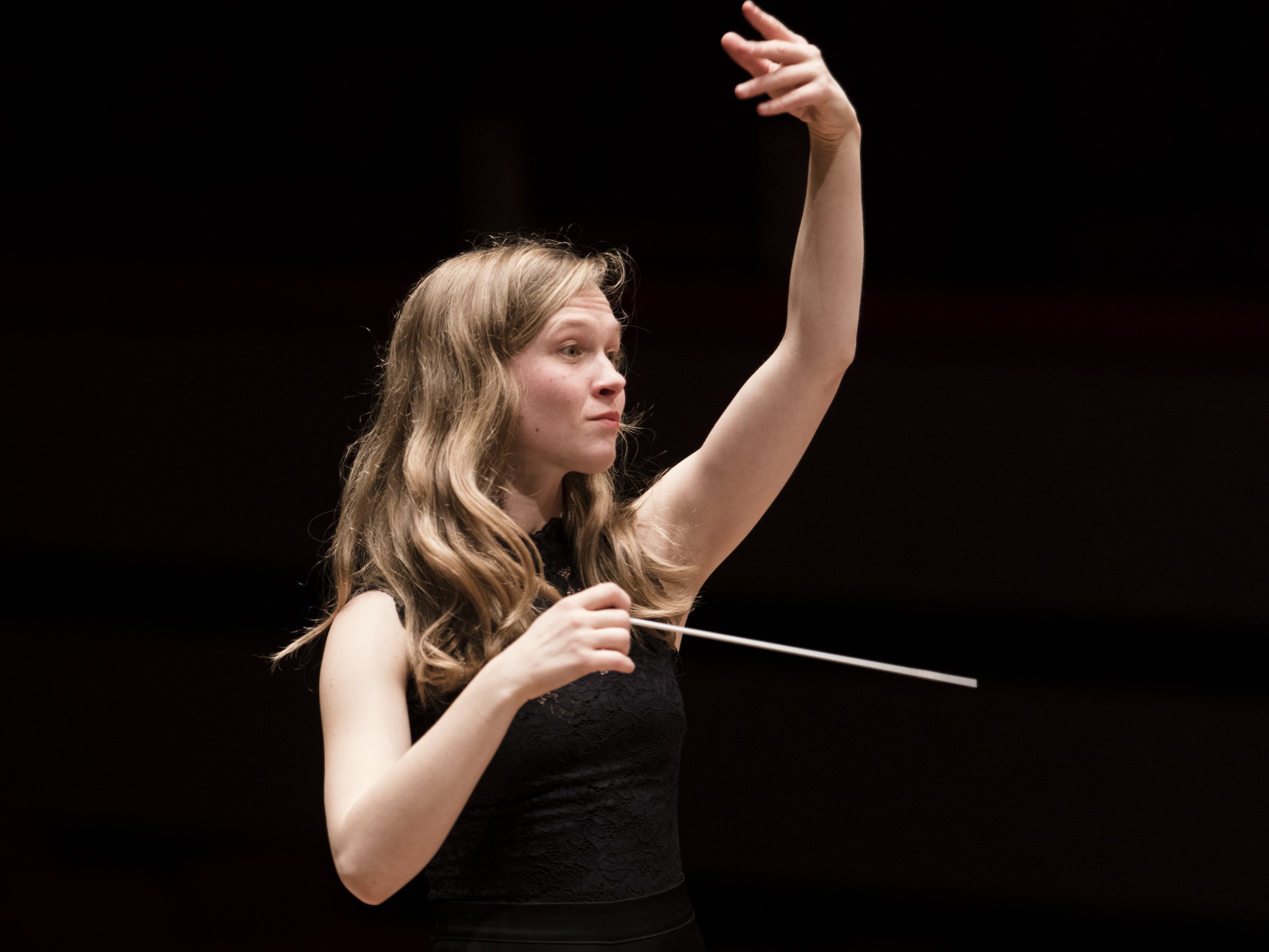 Eine Frau mit langen Haaren dirigiert ein Orchester, hält einen Taktstock in einer Hand und hebt die andere Hand. Sie trägt ein ärmelloses schwarzes Oberteil vor einem dunklen Hintergrund.