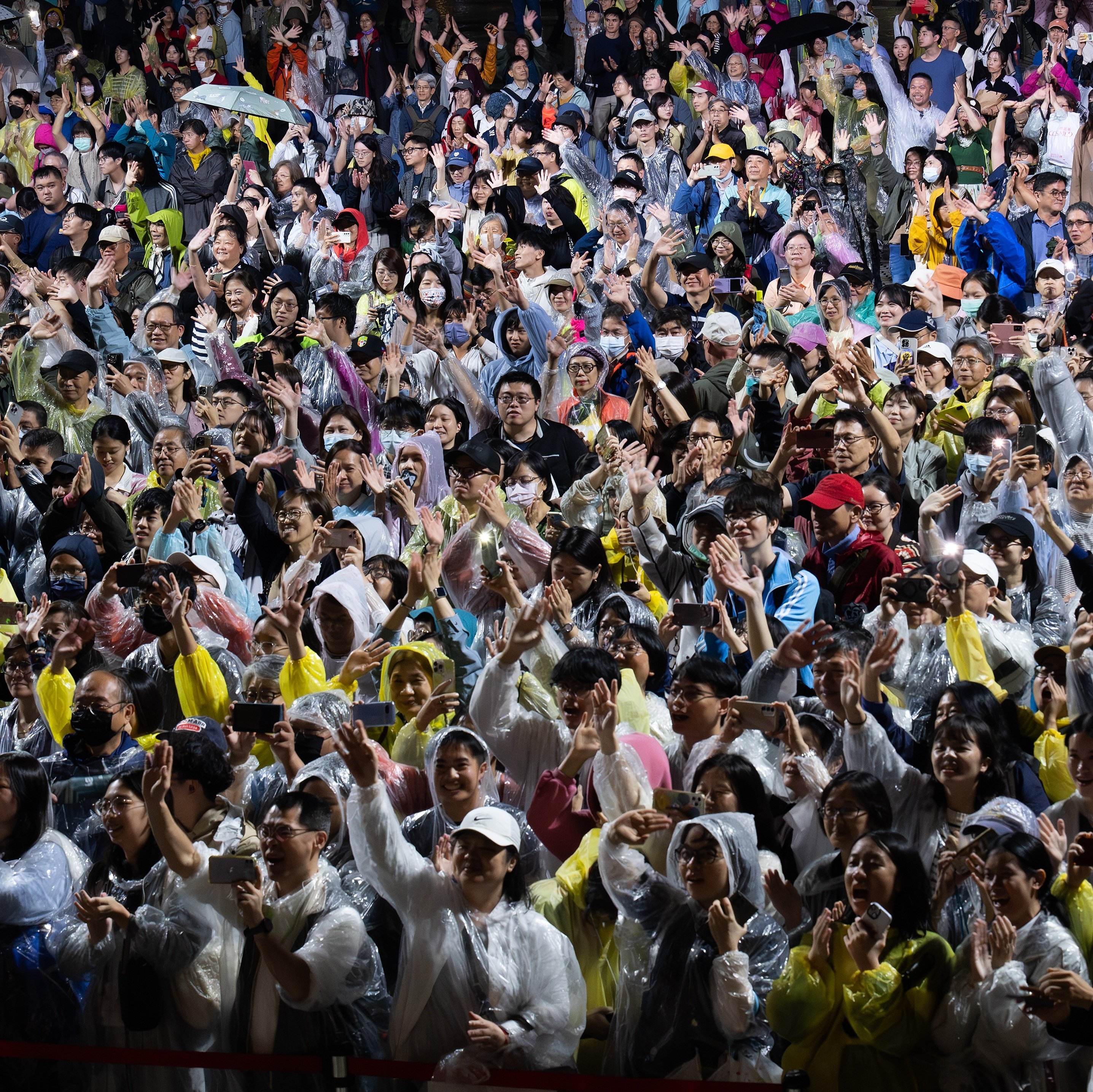 Eine große Menschenmenge in Regenmänteln und Ponchos klatscht und jubelt bei einer nächtlichen Veranstaltung im Freien. Viele Menschen lächeln und heben begeistert die Hände.