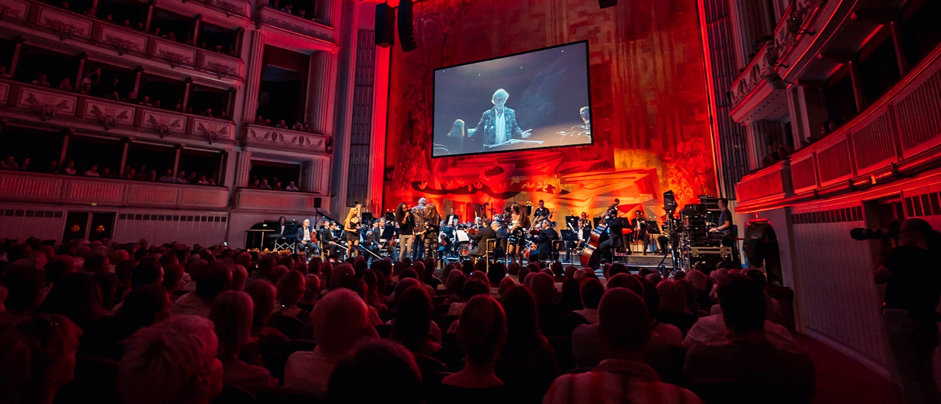 Ein Sinfonieorchester spielt auf der Bühne eines prächtigen Theaters, das in rotes Licht getaucht ist. Auf einer großen Leinwand über den Musikern ist eine Nahaufnahme des Dirigenten zu sehen. Das Publikum füllt die Ränge und verfolgt das Geschehen gebannt.