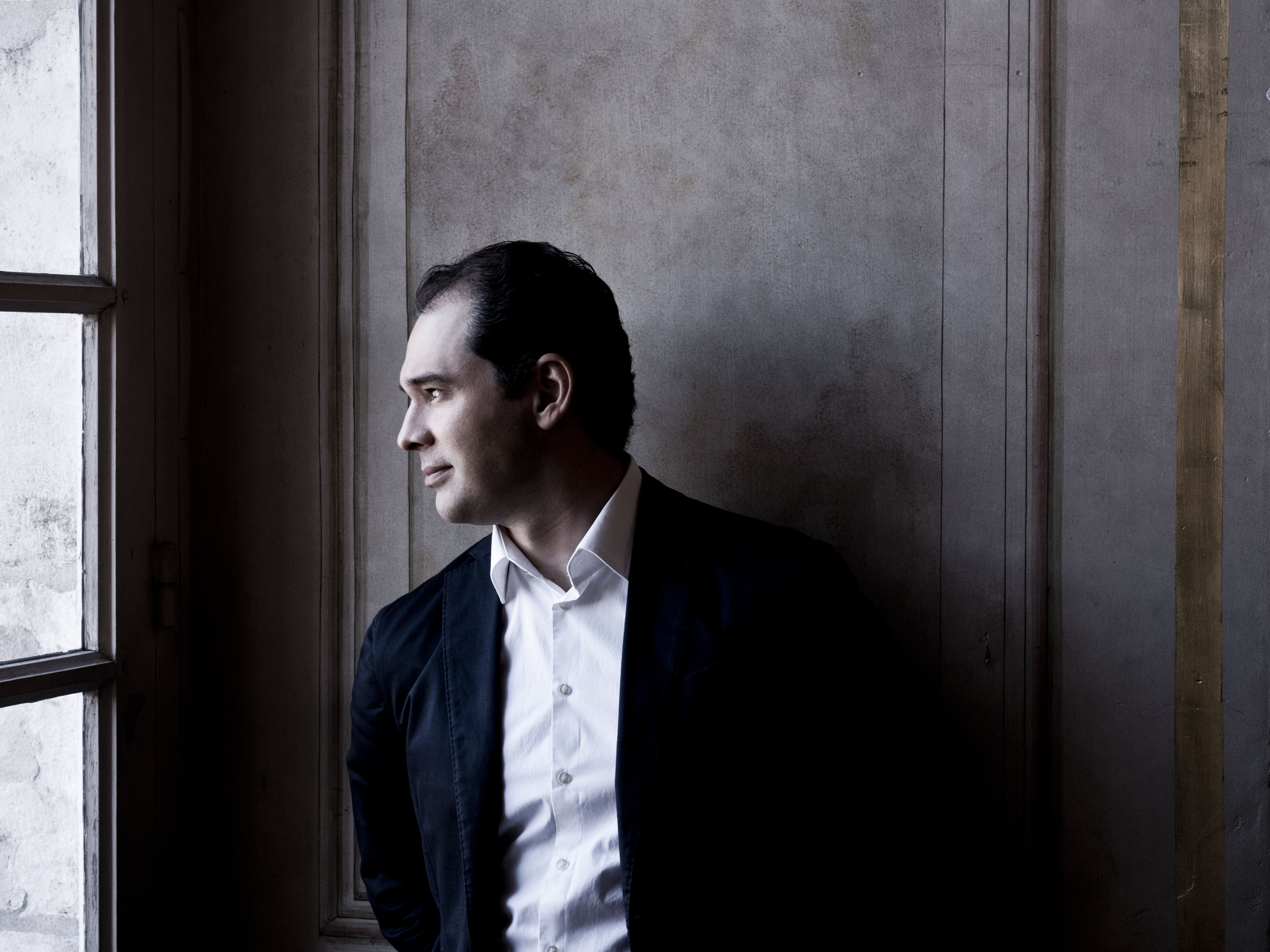 Porträt von Tugan Sokhiev im Anzug und weißen Hemd. Er steht vor einer holzvertäfelten Wand und schaurt links aus einem hohen Fenster.