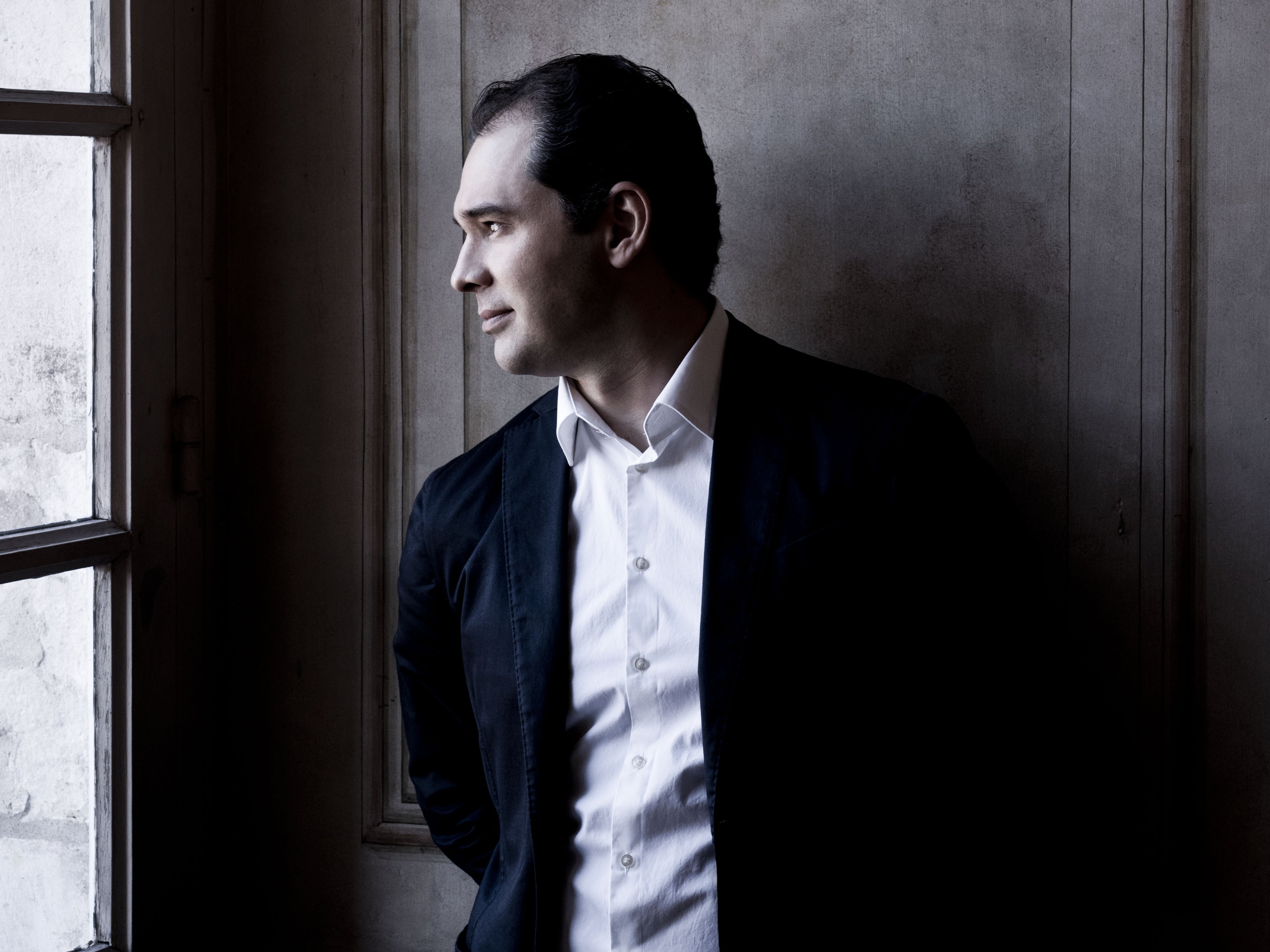 Porträt von Tugan Sokhiev im Anzug und weißen Hemd. Er steht vor einer holzvertäfelten Wand und schaurt links aus einem hohen Fenster.