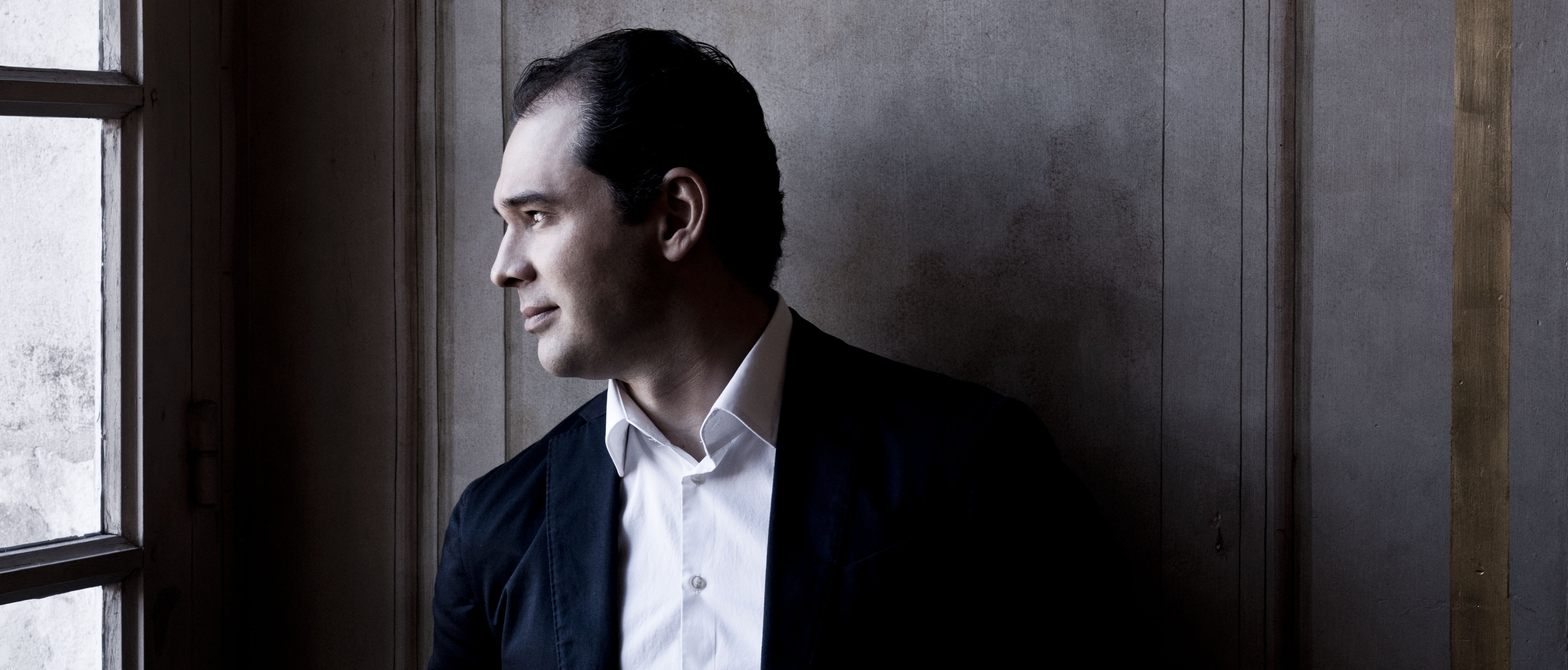 Porträt von Tugan Sokhiev im Anzug und weißen Hemd. Er steht vor einer holzvertäfelten Wand und schaurt links aus einem hohen Fenster.