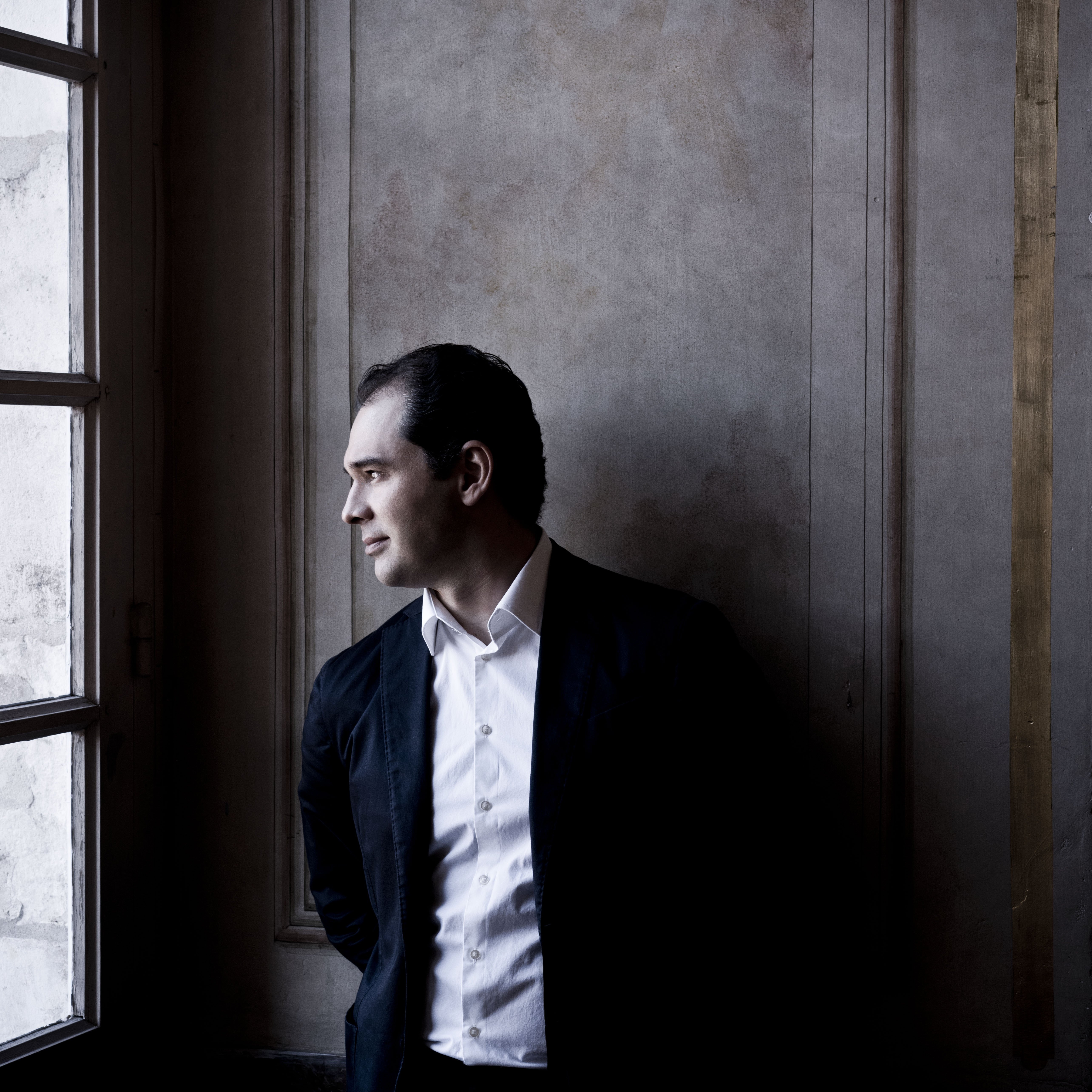 Porträt von Tugan Sokhiev im Anzug und weißen Hemd. Er steht vor einer holzvertäfelten Wand und schaurt links aus einem hohen Fenster.