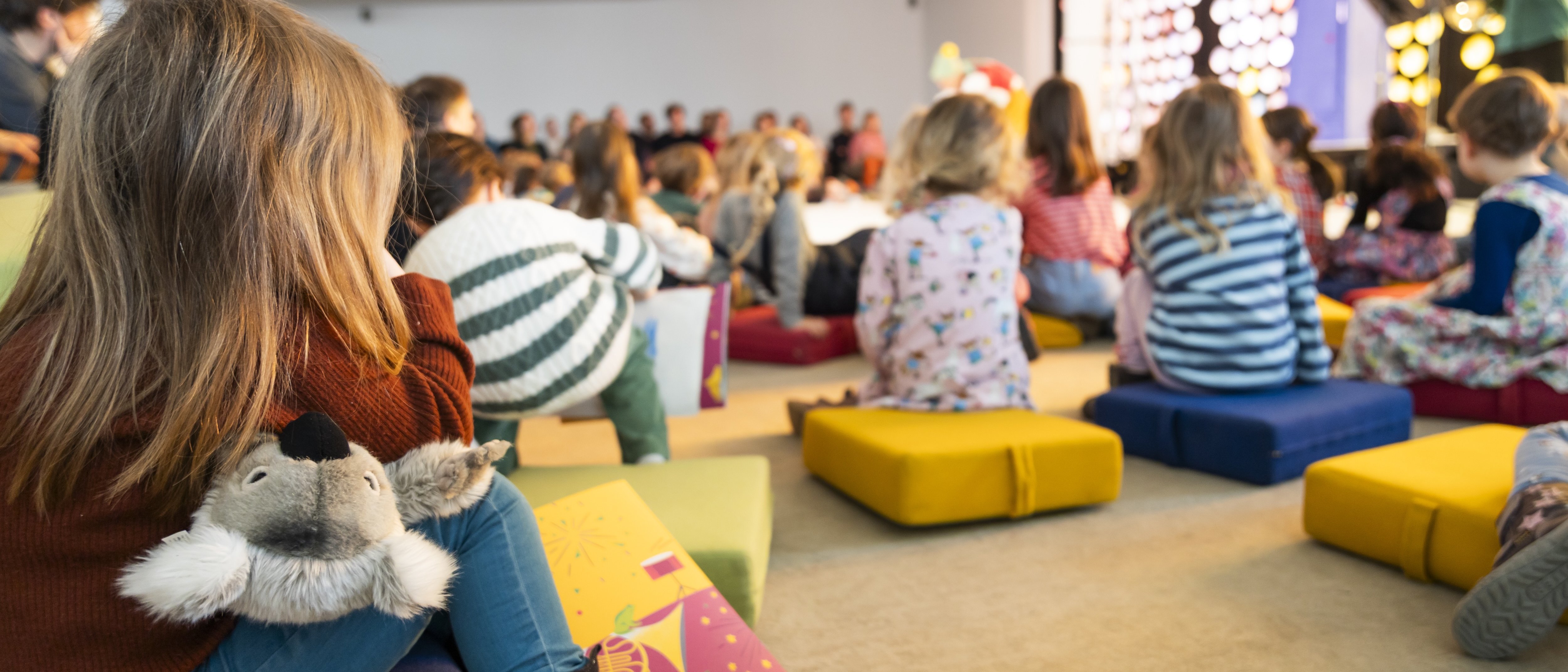 Kinder sitzen auf bunten Kissen vor einer hell erleuchteten Bühne und sehen sich eine Aufführung an. Ein Kind im Vordergrund umarmt ein Stofftier. Die Atmosphäre ist lebhaft und fröhlich.