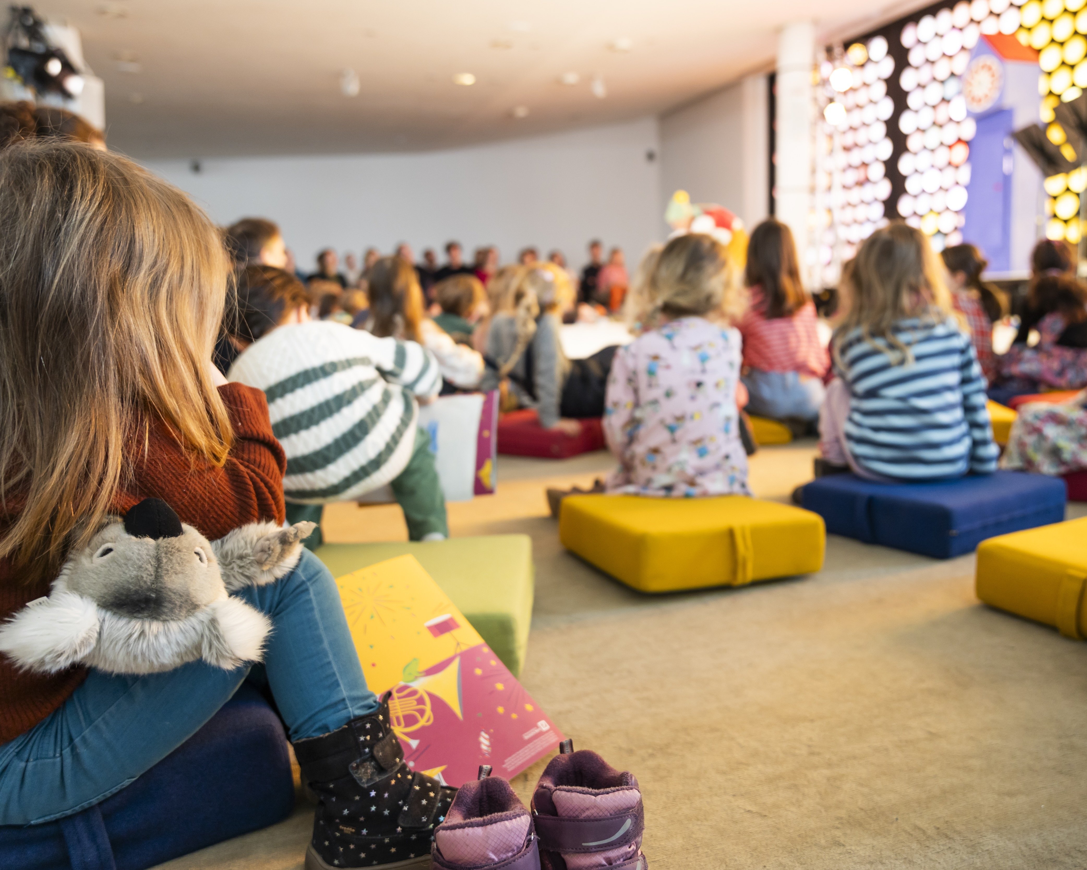 Kinder sitzen auf bunten Kissen vor einer hell erleuchteten Bühne und sehen sich eine Aufführung an. Ein Kind im Vordergrund umarmt ein Stofftier. Die Atmosphäre ist lebhaft und fröhlich.