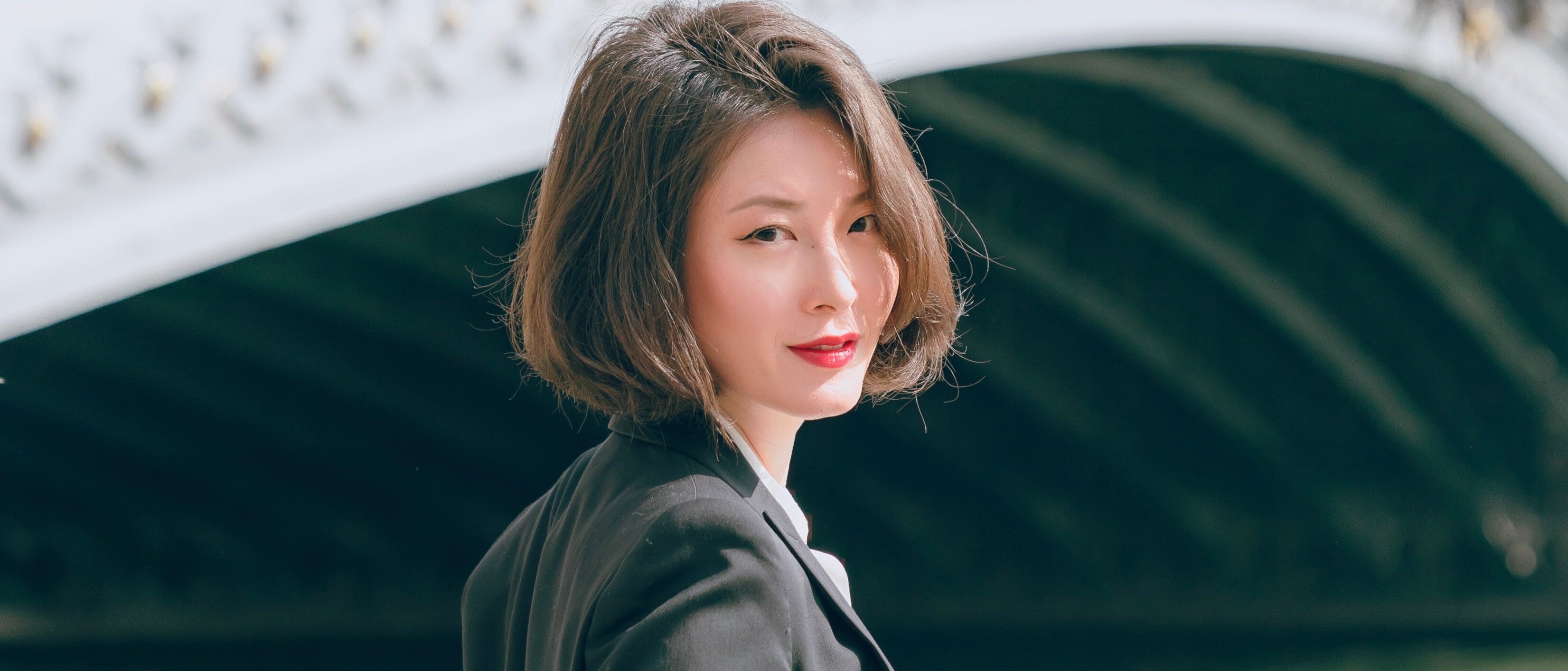 Shin Young Lee mit kurzen braunen Haaren und einem schwarzen Blazer steht leicht lächelnd an einem Fluss, mit einer dekorativen Bogenbrücke im Hintergrund.