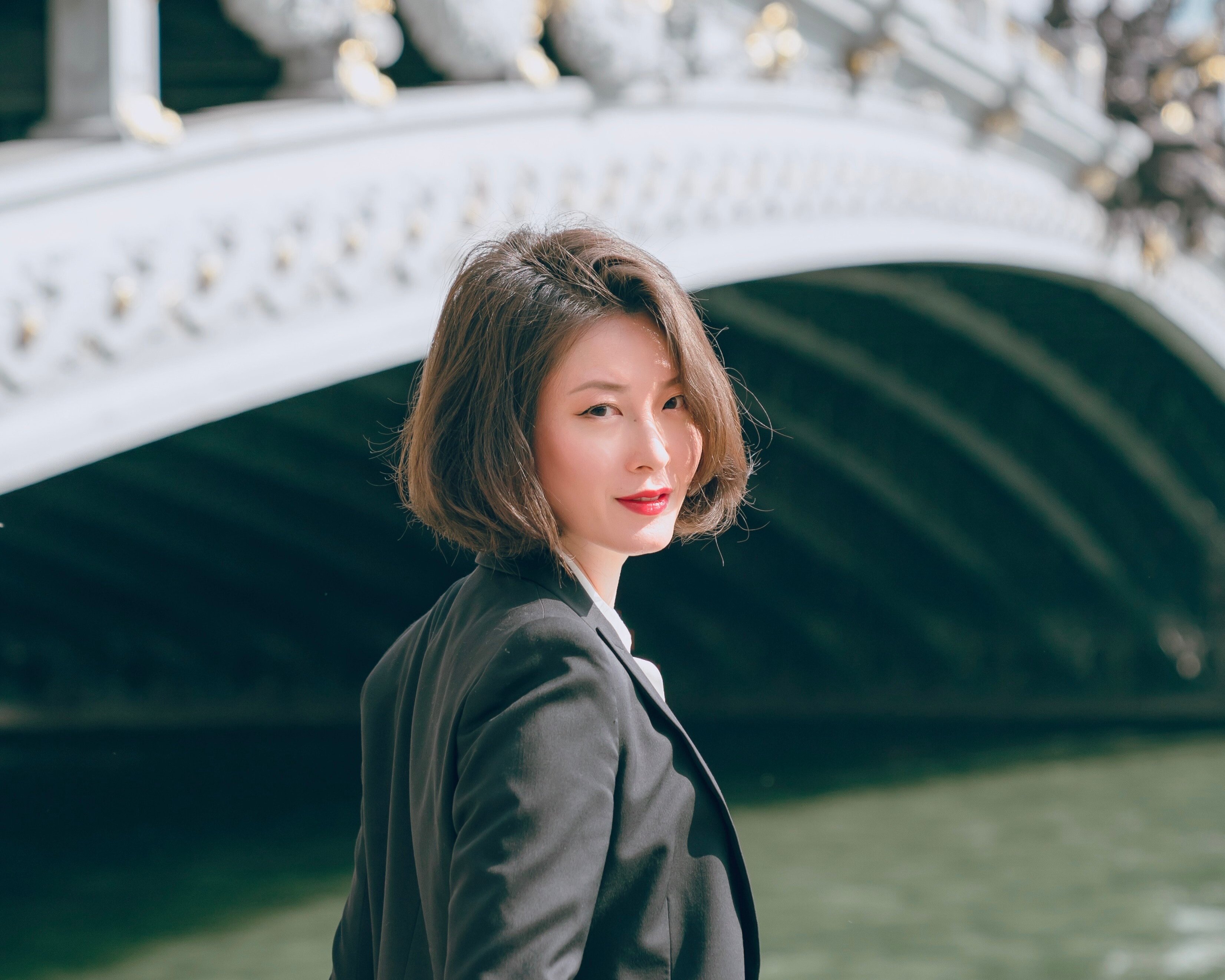 Shin Young Lee mit kurzen braunen Haaren und einem schwarzen Blazer steht leicht lächelnd an einem Fluss, mit einer dekorativen Bogenbrücke im Hintergrund.