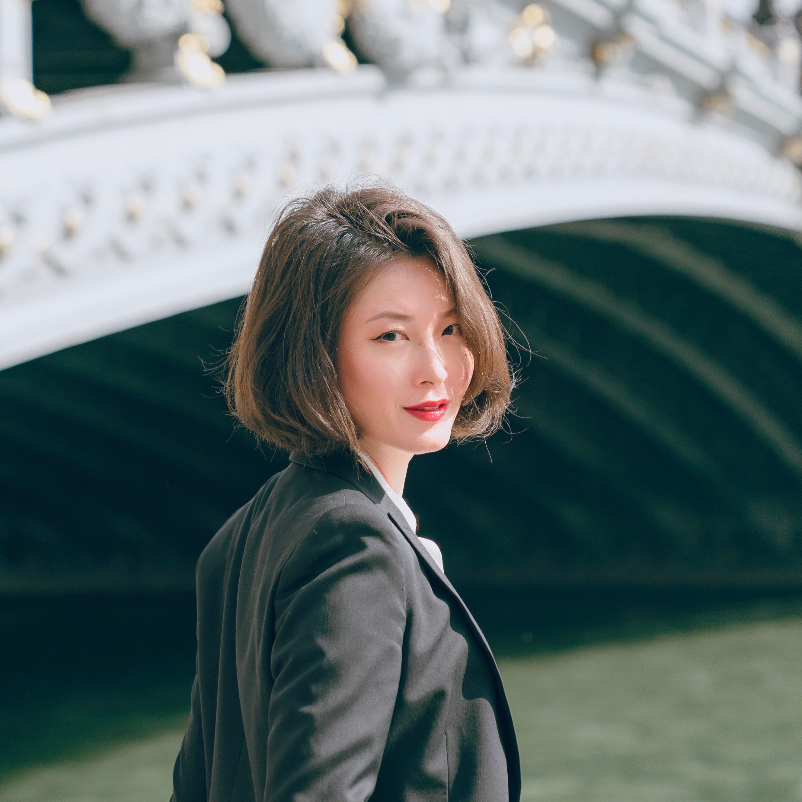 Shin Young Lee mit kurzen braunen Haaren und einem schwarzen Blazer steht leicht lächelnd an einem Fluss, mit einer dekorativen Bogenbrücke im Hintergrund.