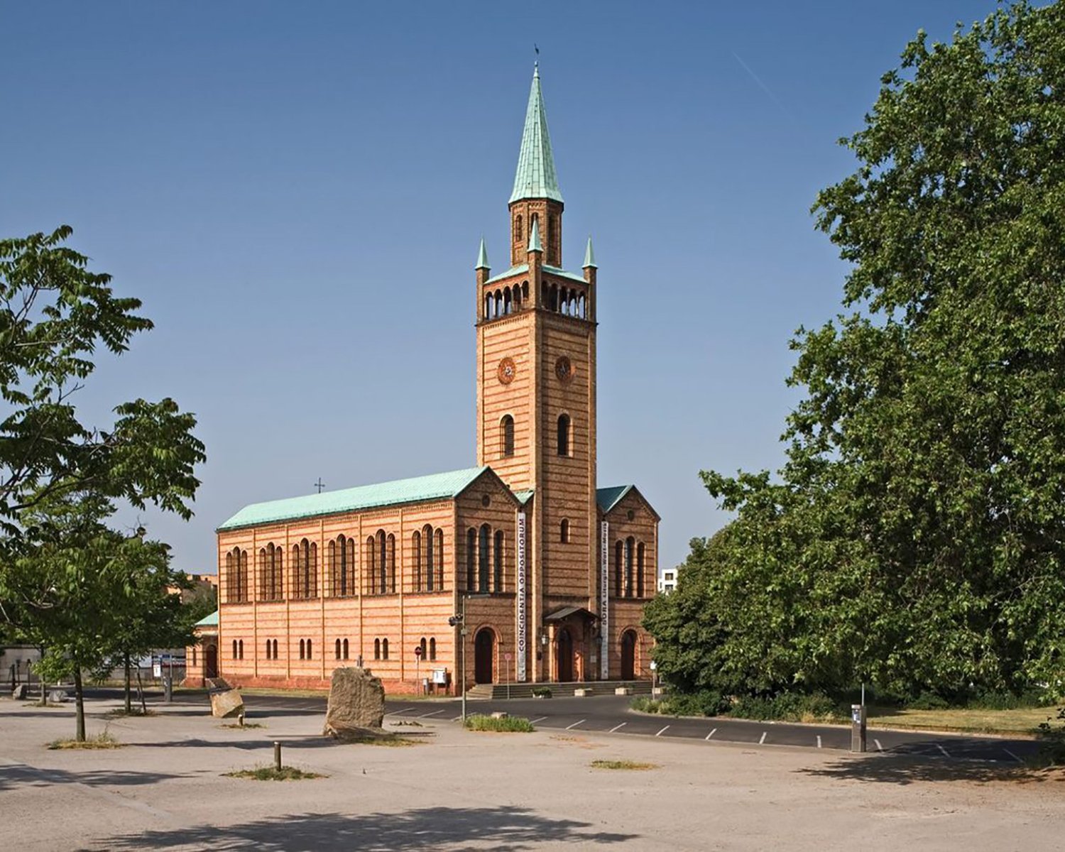 Eine Backsteinkirche mit einem hohen, spitzen grünen Kirchturm steht auf einem offenen Platz mit Bäumen und einem größtenteils leeren Parkplatz unter einem klaren blauen Himmel.