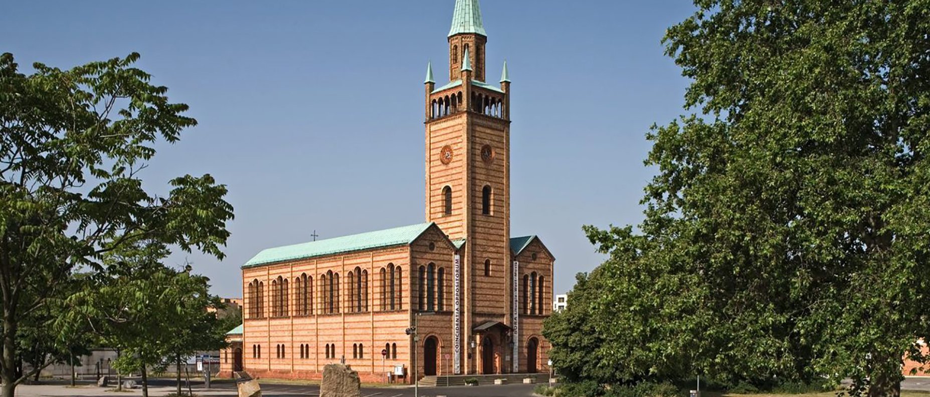 Eine Backsteinkirche mit einem hohen, spitzen grünen Kirchturm steht auf einem offenen Platz mit Bäumen und einem größtenteils leeren Parkplatz unter einem klaren blauen Himmel.
