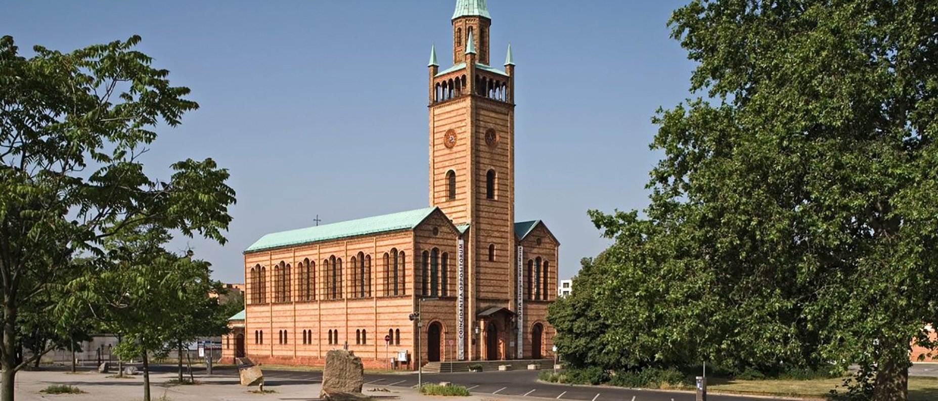 Eine Backsteinkirche mit einem hohen, spitzen grünen Kirchturm steht auf einem offenen Platz mit Bäumen und einem größtenteils leeren Parkplatz unter einem klaren blauen Himmel.