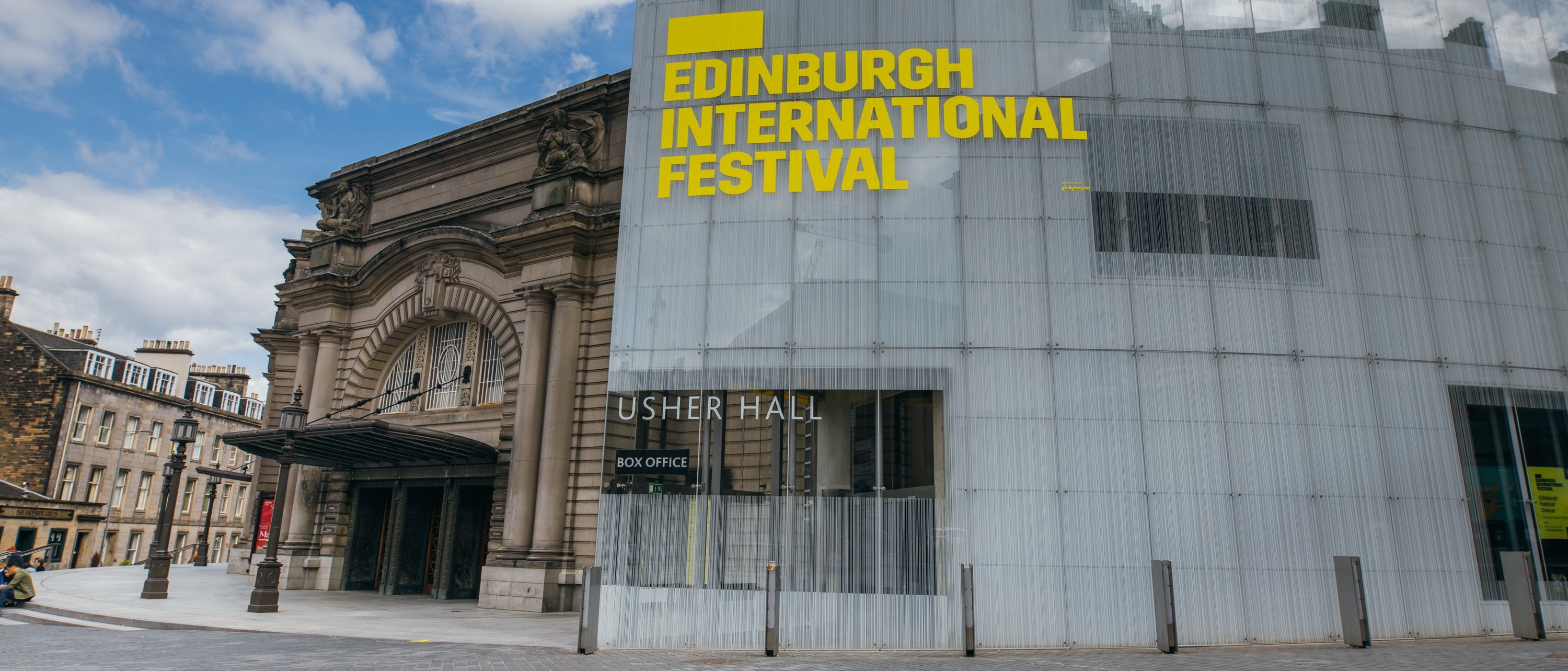 Ein modernes Glasgebäude mit der Aufschrift Edinburgh International Festival in fetten gelben Buchstaben steht neben der historischen Usher Hall unter einem blauen Wolkenhimmel.