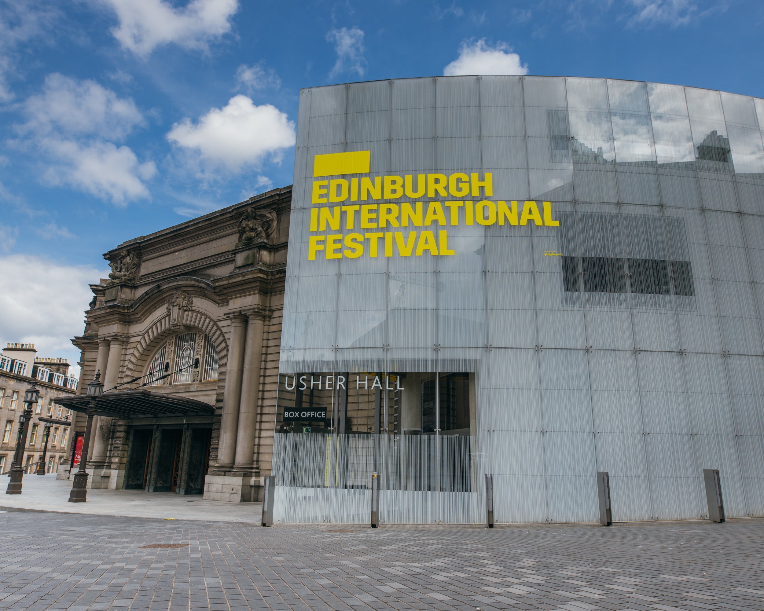 Modern glass-fronted building with bold yellow Edinburgh International Festival signage next to the historic stone Usher Hall, under a partly cloudy blue sky. People and ticket office are visible.