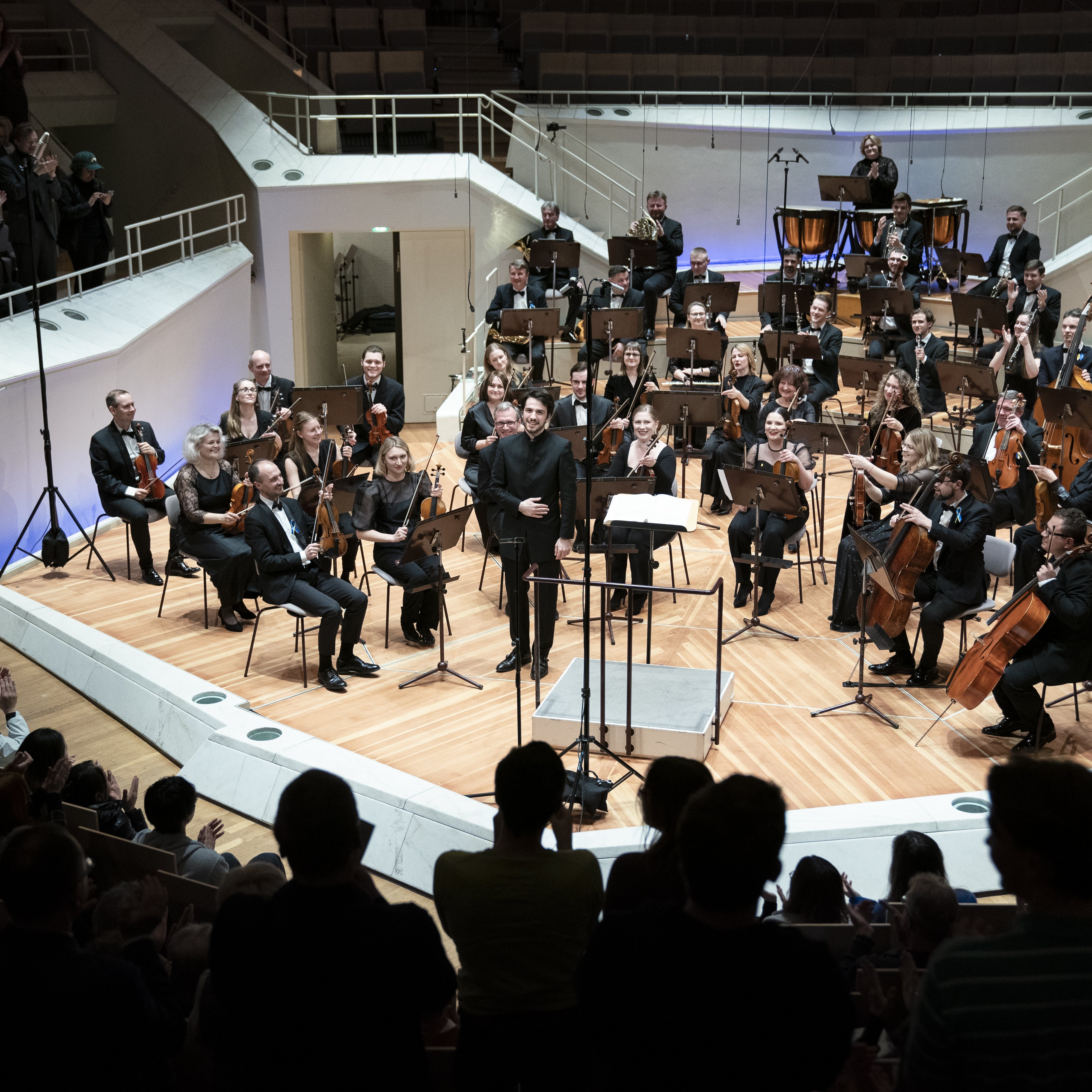 Das Kyiv Symphony Orchestra sitzt auf der Bühne beim Schlussapplaus. Der Dirigent Christian Blex verbeugt sich. Im Vordergrund sind klatschende Menschen zu sehen.