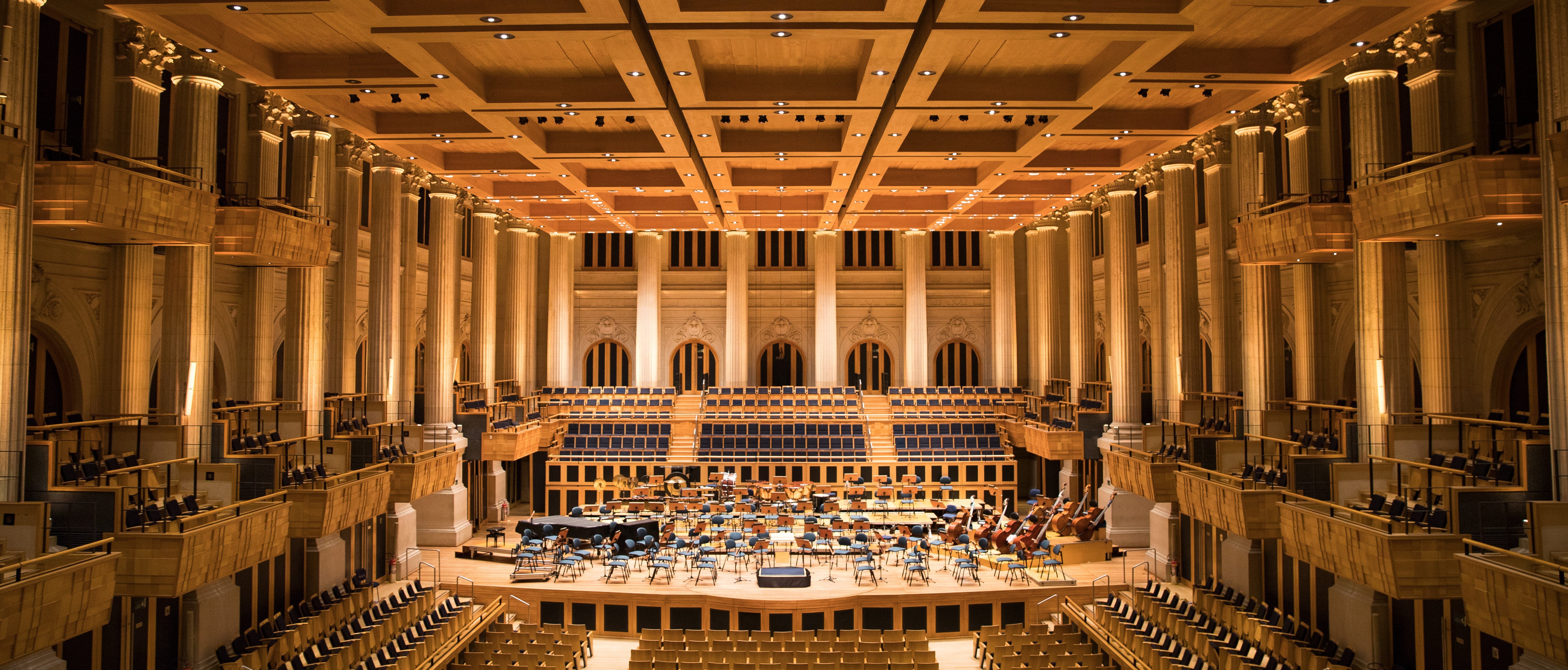 A grand, empty concert hall with rows of seats, a wooden stage set for an orchestra, and warm lighting highlighting the architectural details and high ceiling.