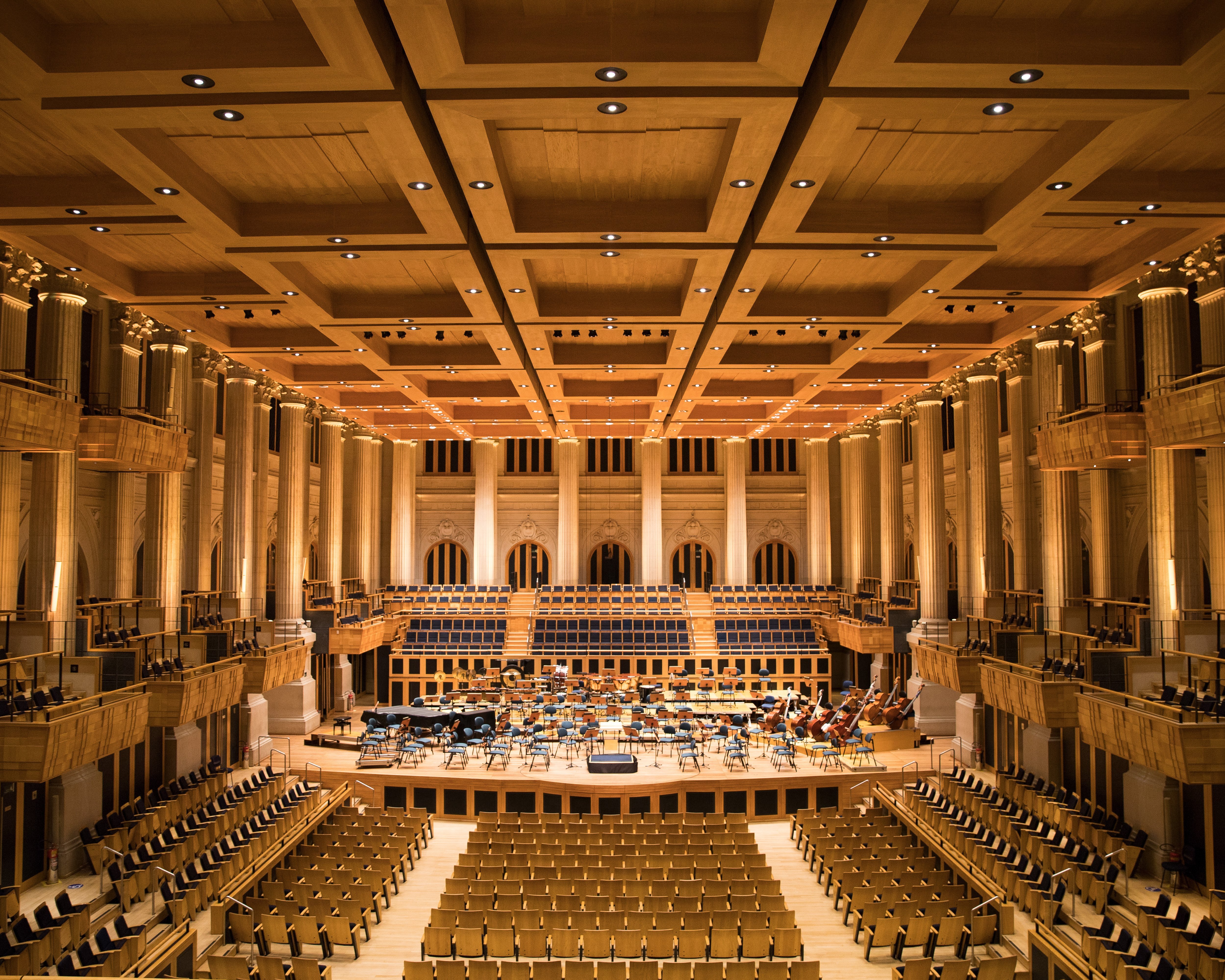 A grand, empty concert hall with rows of seats, a wooden stage set for an orchestra, and warm lighting highlighting the architectural details and high ceiling.