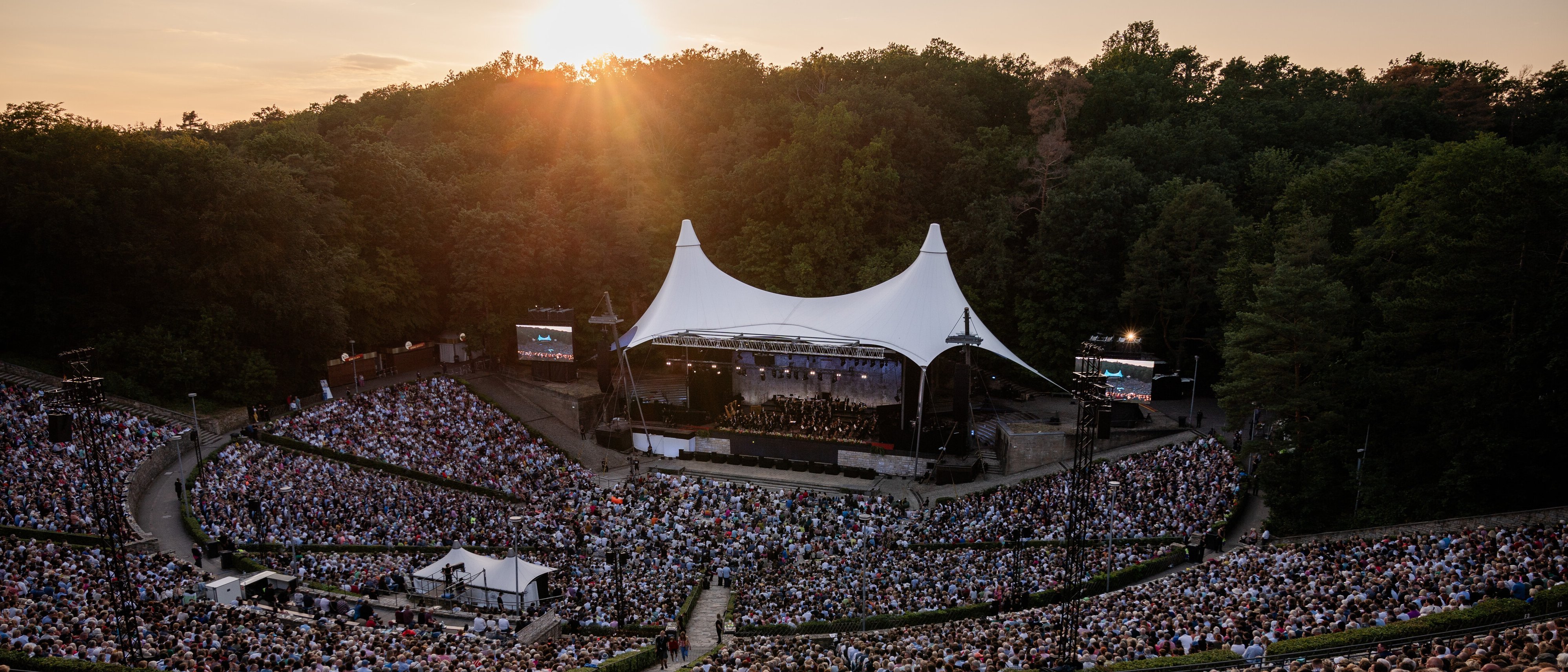 Eine große Menschenmenge verfolgt ein Freiluftkonzert bei Sonnenuntergang in einem von Bäumen umgebenen Amphitheater, in dem ein weißes Zelt die Bühne bedeckt und Musiker auftreten.