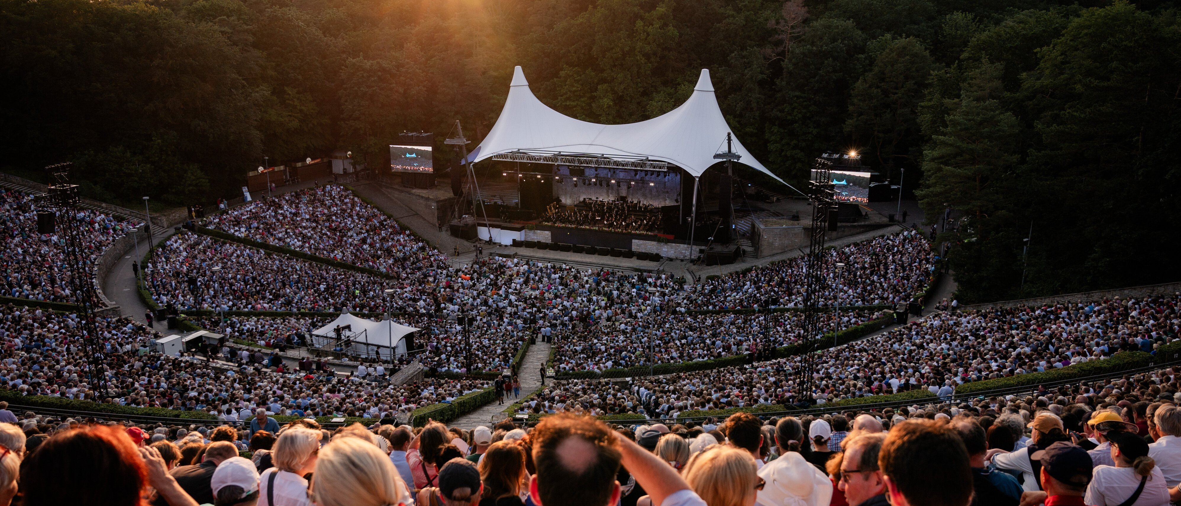 A large outdoor concert with a white tented stage, surrounded by a big crowd seated in an amphitheater, all under the setting sun and a sky tinged with warm orange hues, bordered by trees.