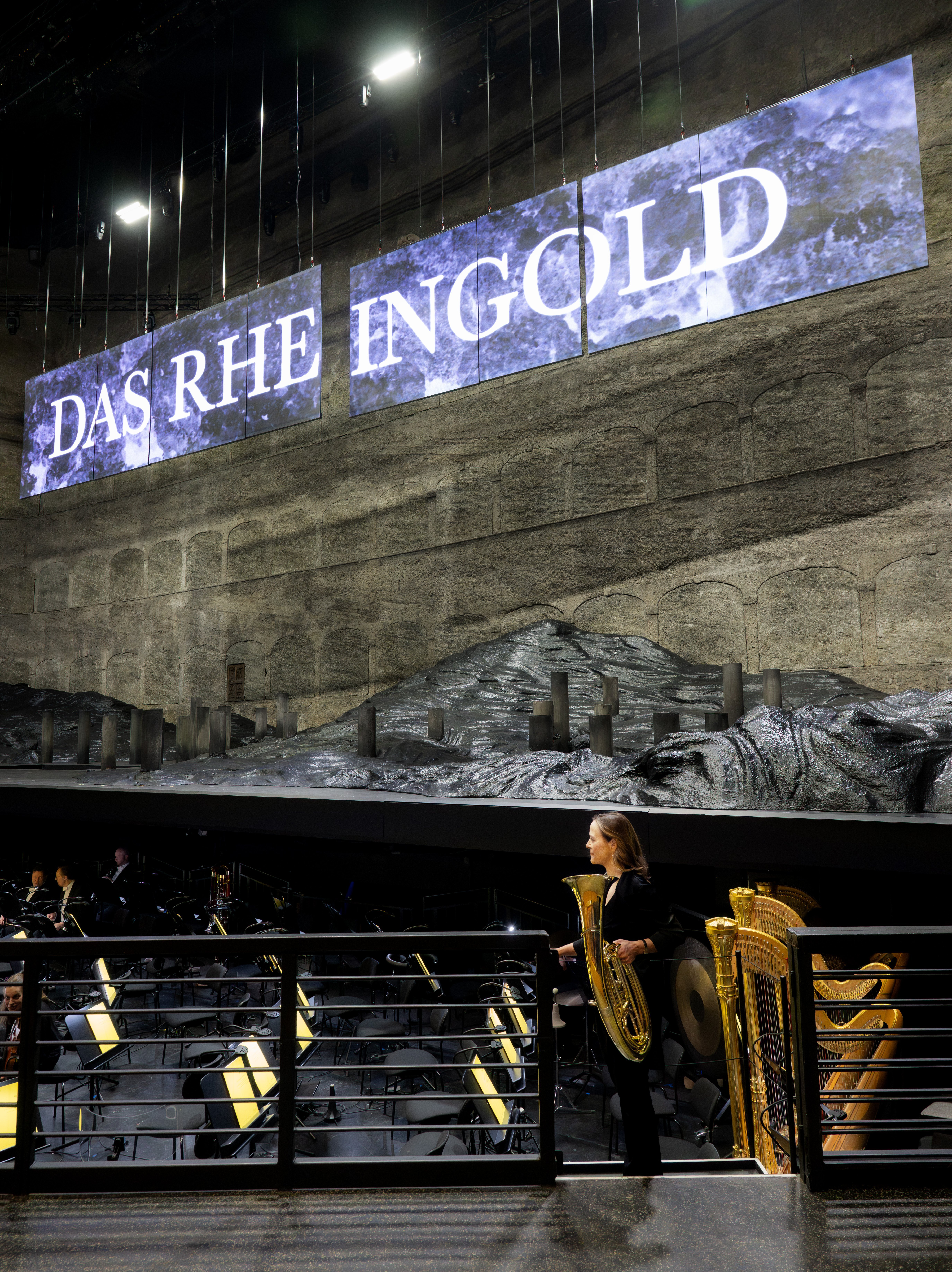 Hornistin Sarah Willis betritt den Orchestergraben vor der Vorstellung, im Hintergrund sieht man die Bühne, sowie Bildschirme mit der Aufschrift »Das Rheingold«.