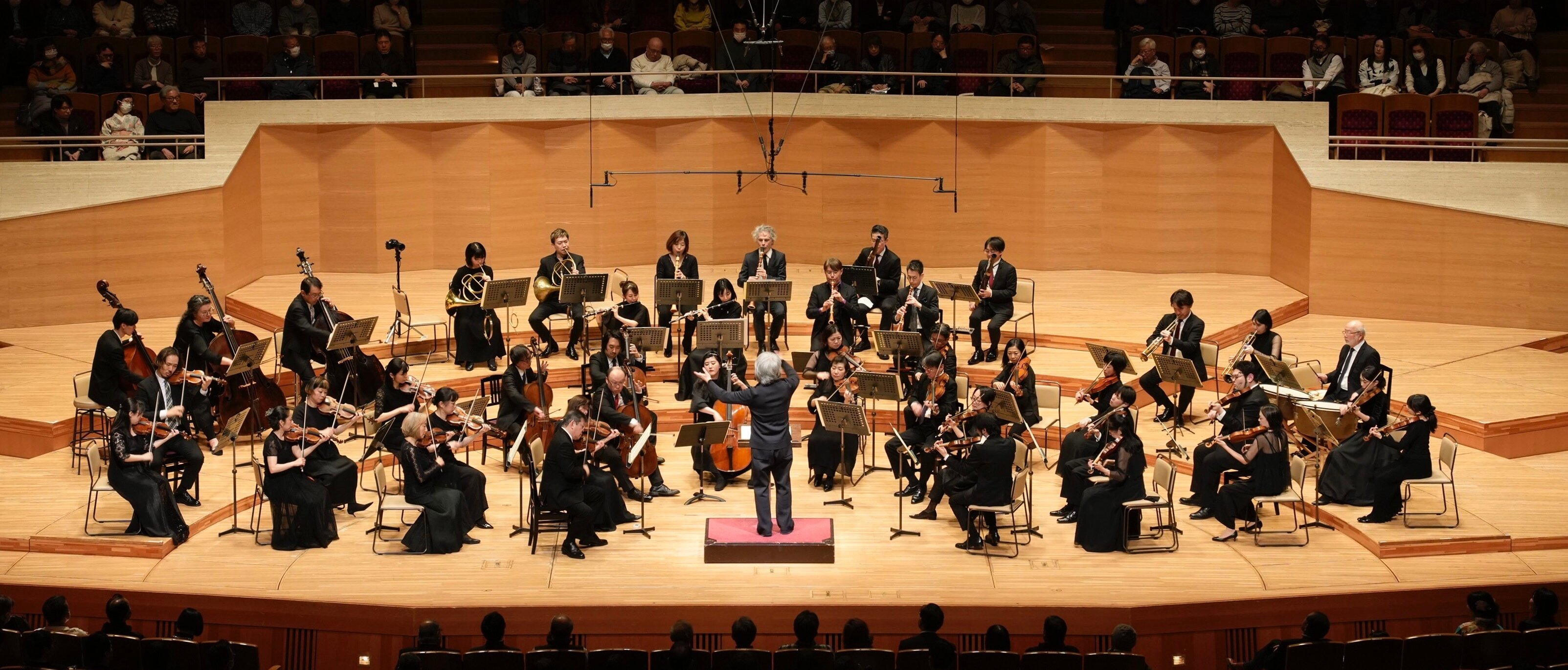 Ein Dirigent leitet ein Orchester auf einer gut beleuchteten Bühne in einem Konzertsaal, während die Musiker verschiedene Instrumente spielen und das Publikum im Hintergrund sitzt.