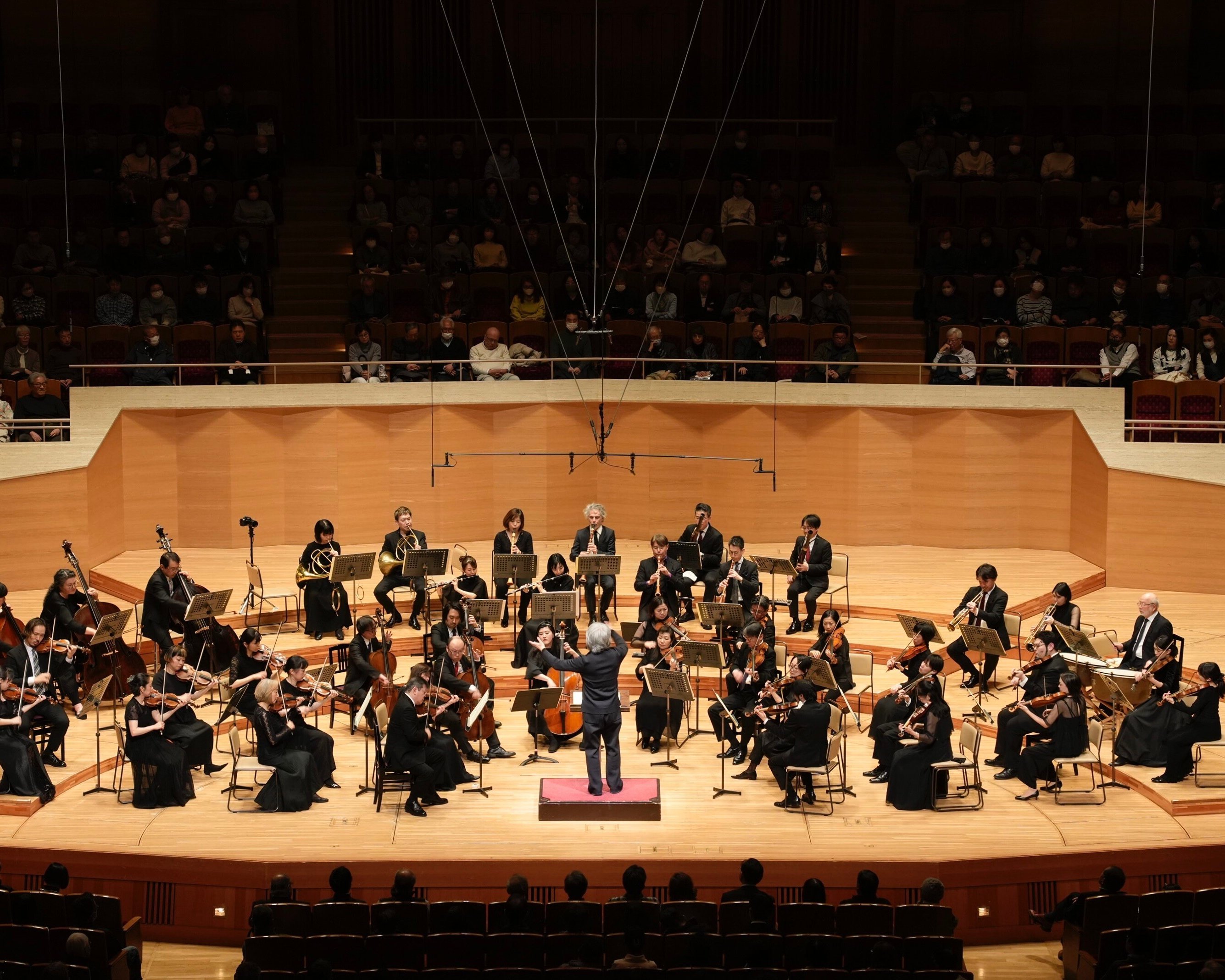 Ein Dirigent leitet ein Orchester auf einer gut beleuchteten Bühne in einem Konzertsaal, während die Musiker verschiedene Instrumente spielen und das Publikum im Hintergrund sitzt.