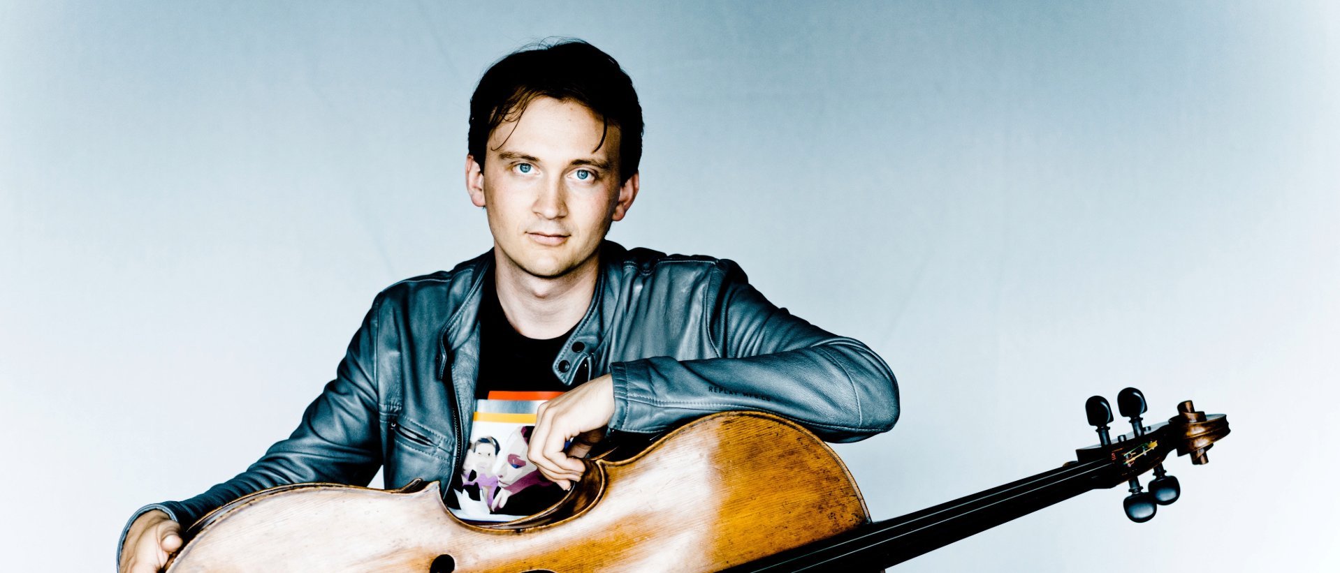Ein junger Mann in einer grauen Jacke sitzt da, hält ein Cello in den Händen und stützt seinen Arm auf das Instrument. Er hat kurzes braunes Haar und einen ausdruckslosen Gesichtsausdruck; hinter ihm ist ein weißer Hintergrund zu sehen.