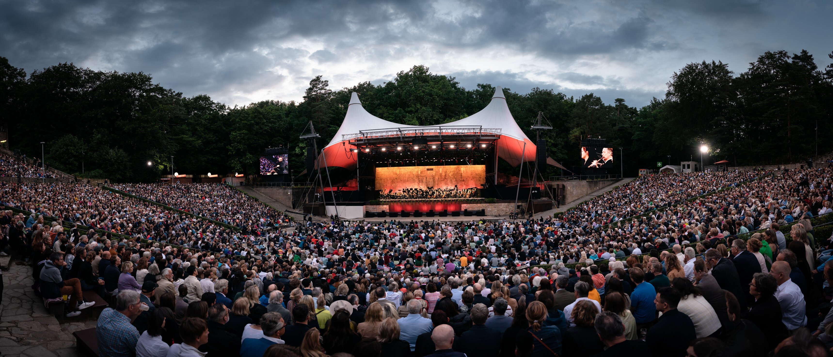 Ein mit Zuschauern gefülltes Amphitheater im Freien steht vor einer hell erleuchteten Bühne mit einem großen Vordach, umgeben von Bäumen unter einem bewölkten Abendhimmel. Auf der Bühne treten Musiker auf.