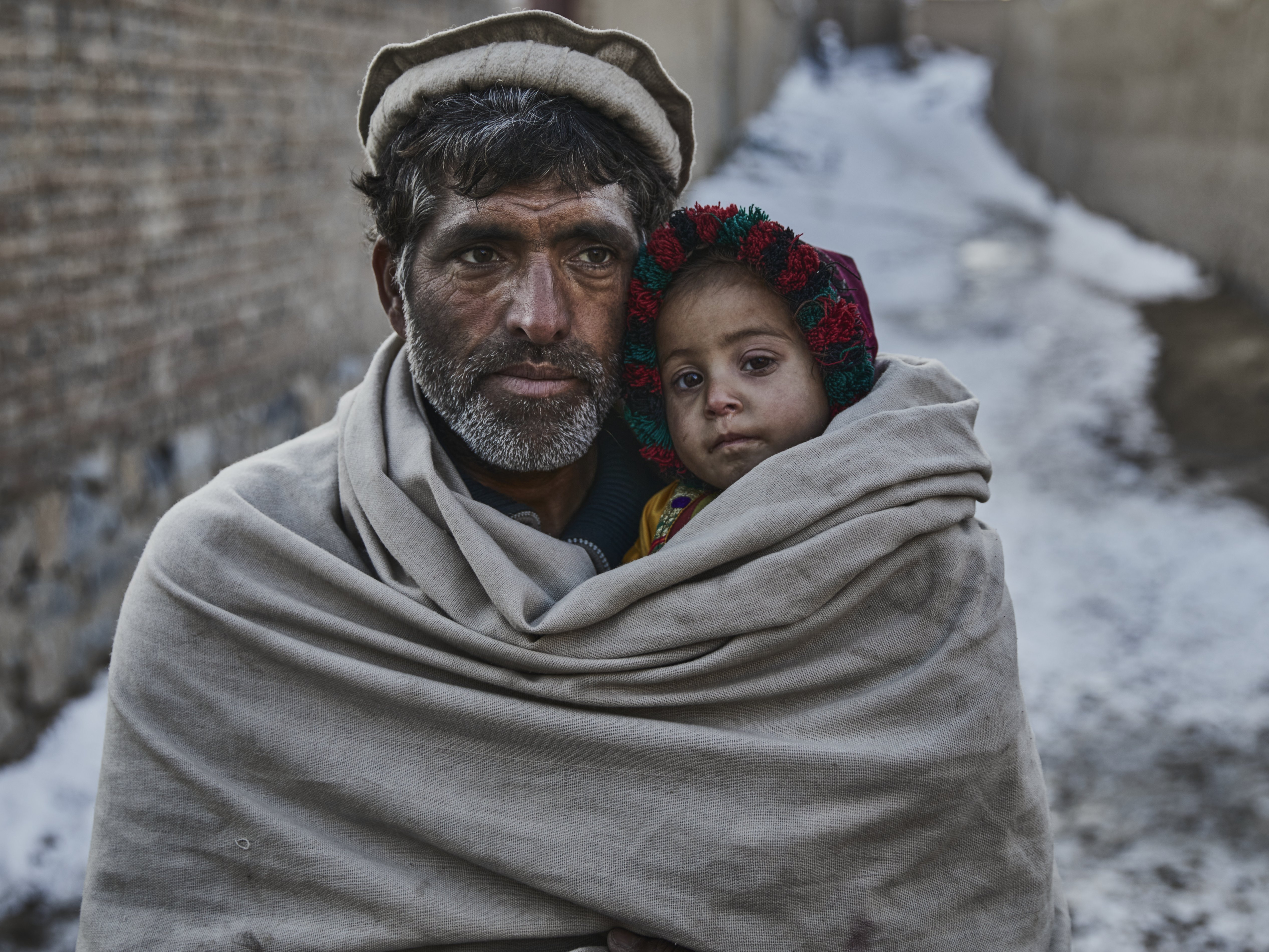 Ein Mann trägt ein kleines Kind in einer wärmenden Decke durch eine schneebedeckte Gasse. Beide haben ernste Gesichter; das Kind trägt eine bunte Mütze. Die Umgebung wirkt kalt und schlicht, mit Mauern aus Stein und Schnee im Hintergrund.