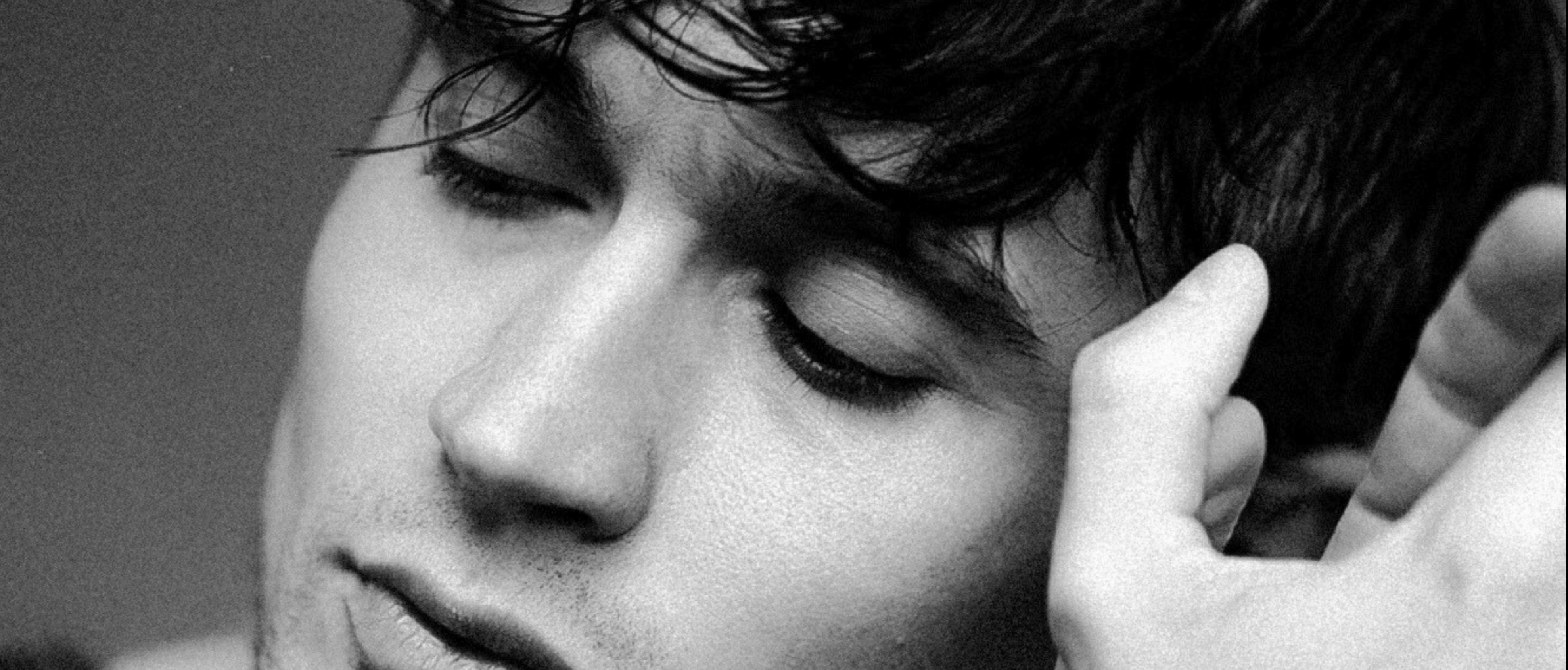 A close-up black and white photo of a young man with tousled dark hair, closed eyes, and a relaxed expression, resting his head on his hand with fingers visible near his face.