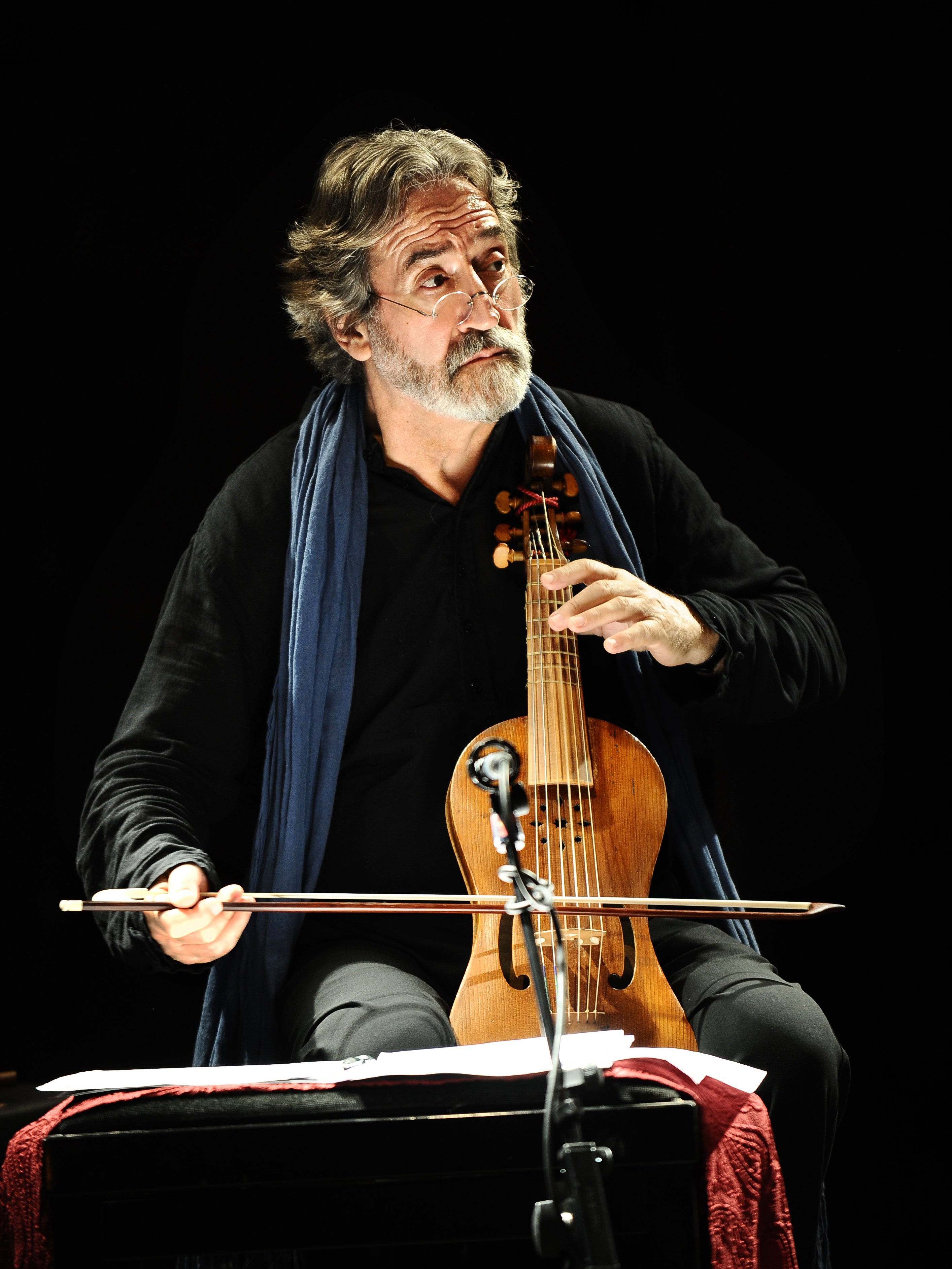 A bearded man with gray hair, wearing a dark shirt and blue scarf, plays a bowed string instrument on stage, focused intently, with sheet music in front of him and a microphone nearby.