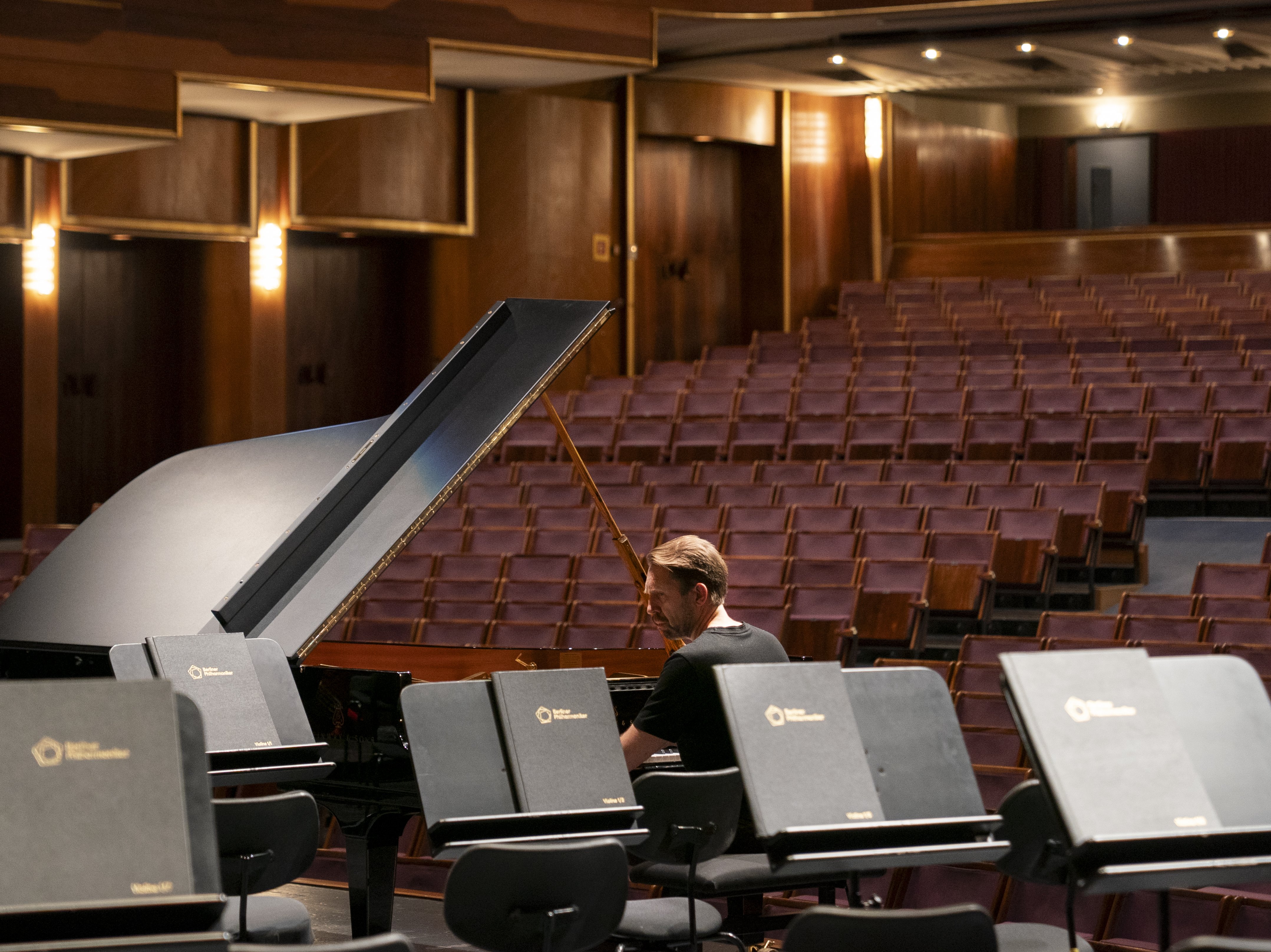 A person plays a grand piano on a stage in an empty concert hall, surrounded by vacant music stands and rows of unoccupied seats under warm lighting.