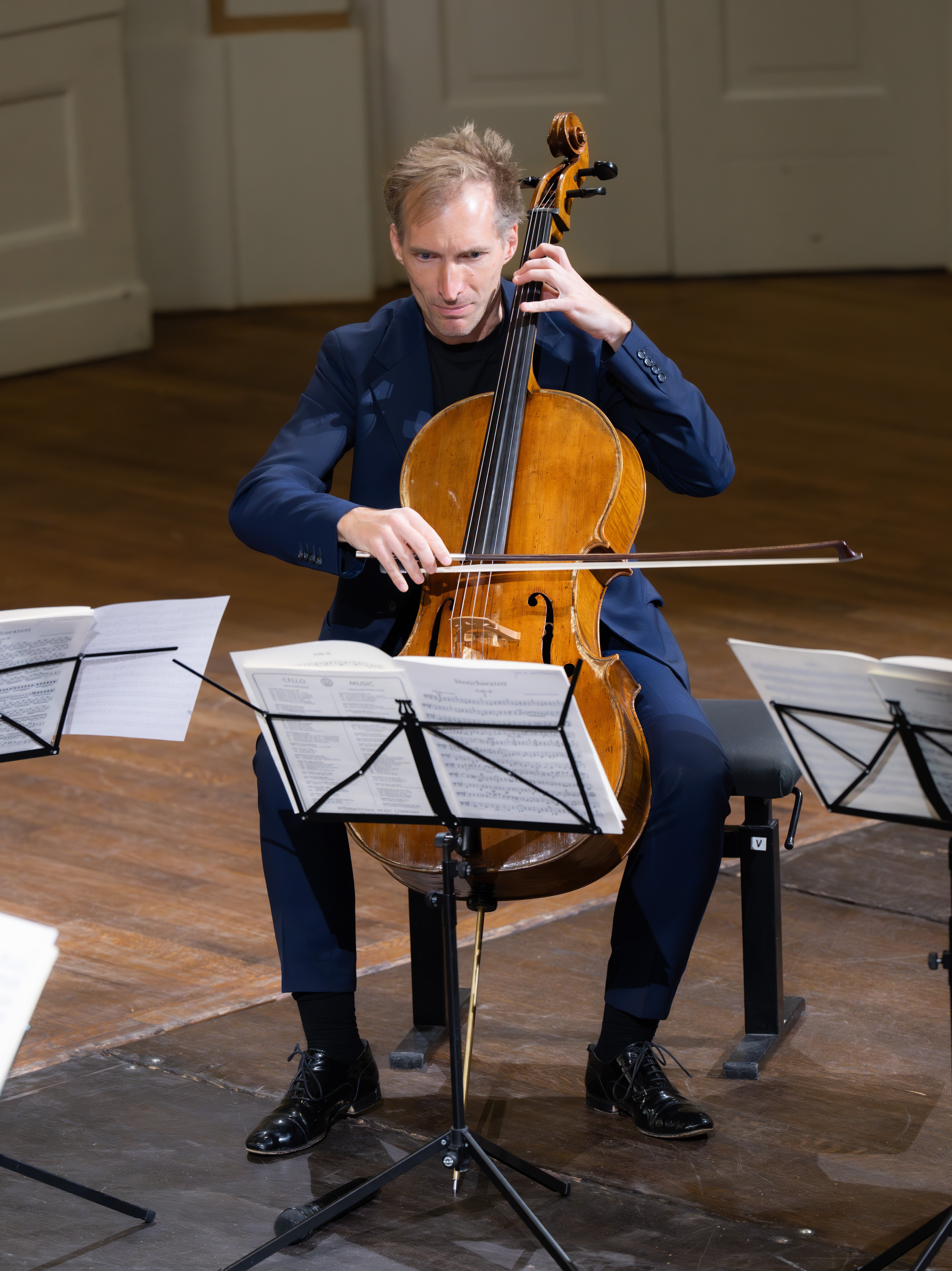 Ein Cellist in einem dunklen Anzug spielt auf der Bühne, umgeben von Notenständern mit Notenblättern, in einem gut beleuchteten Konzertsaal mit Holzfußboden und weißen Wänden Cello.