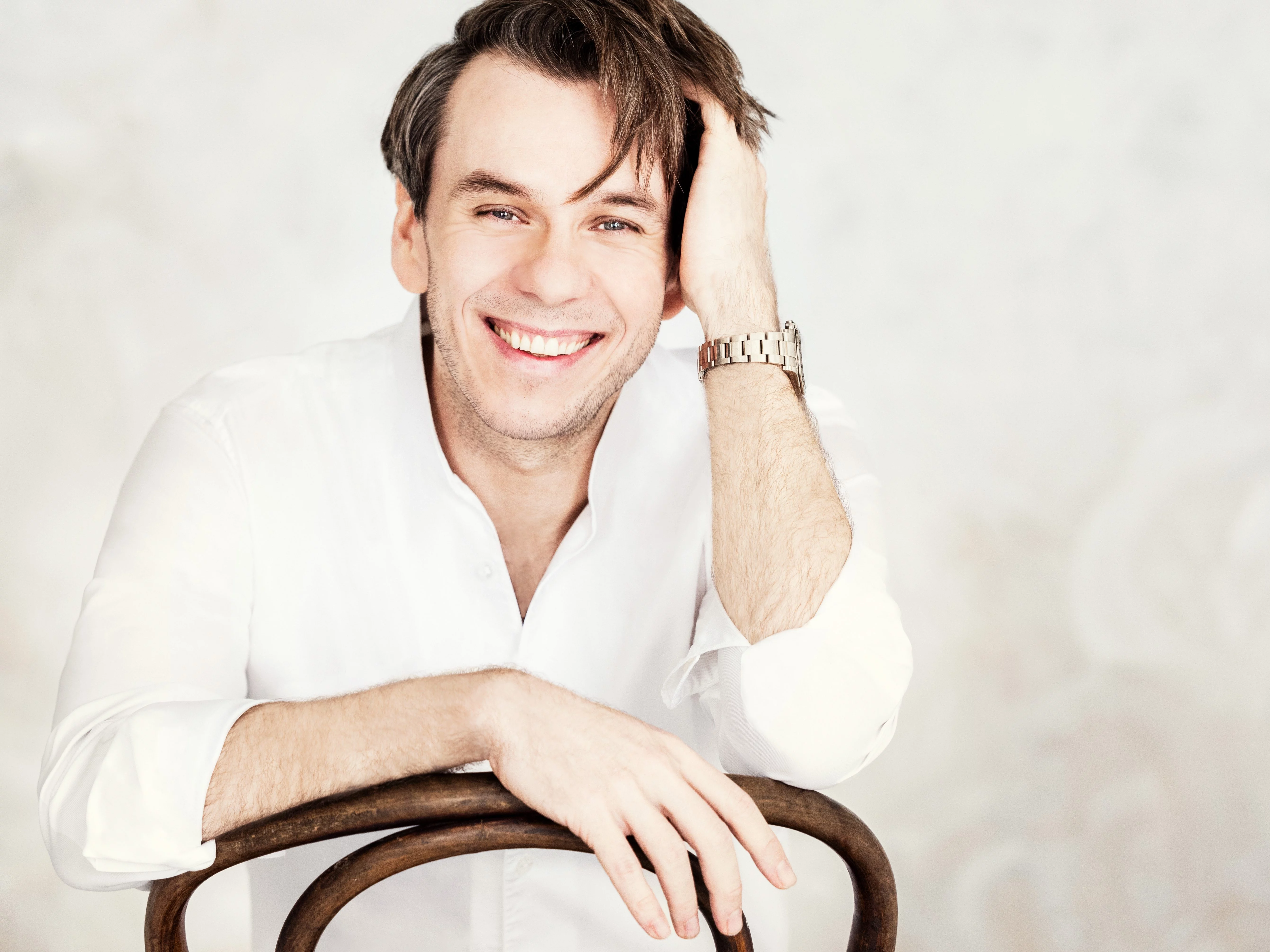 Benjamin Bernheim sitzt lächelnd auf einem Stuhl, trägt ein weißes Hemd und eine Uhr und seine Hand ruht auf dem Kopf, vor einem hellen, strukturierten Hintergrund.