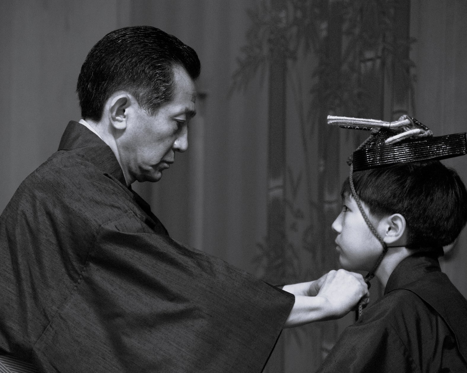 Ein Mann in traditioneller japanischer Kleidung rückt den Kimono einer jungen Person zurecht, die eine verzierte Kopfbedeckung trägt. Sie stehen sich in einem ruhigen Innenraum gegenüber. Das Bild ist in schwarz-weiß gehalten.