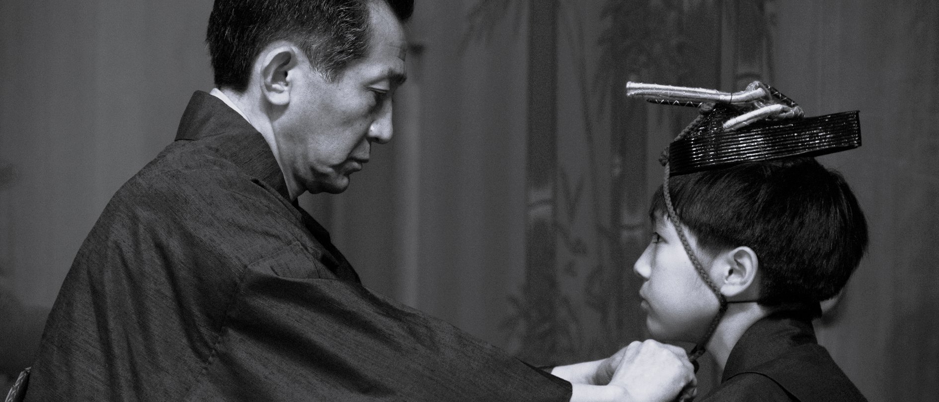 Ein Mann in traditioneller japanischer Kleidung rückt den Kimono einer jungen Person zurecht, die eine verzierte Kopfbedeckung trägt. Sie stehen sich in einem ruhigen Innenraum gegenüber. Das Bild ist in schwarz-weiß gehalten.