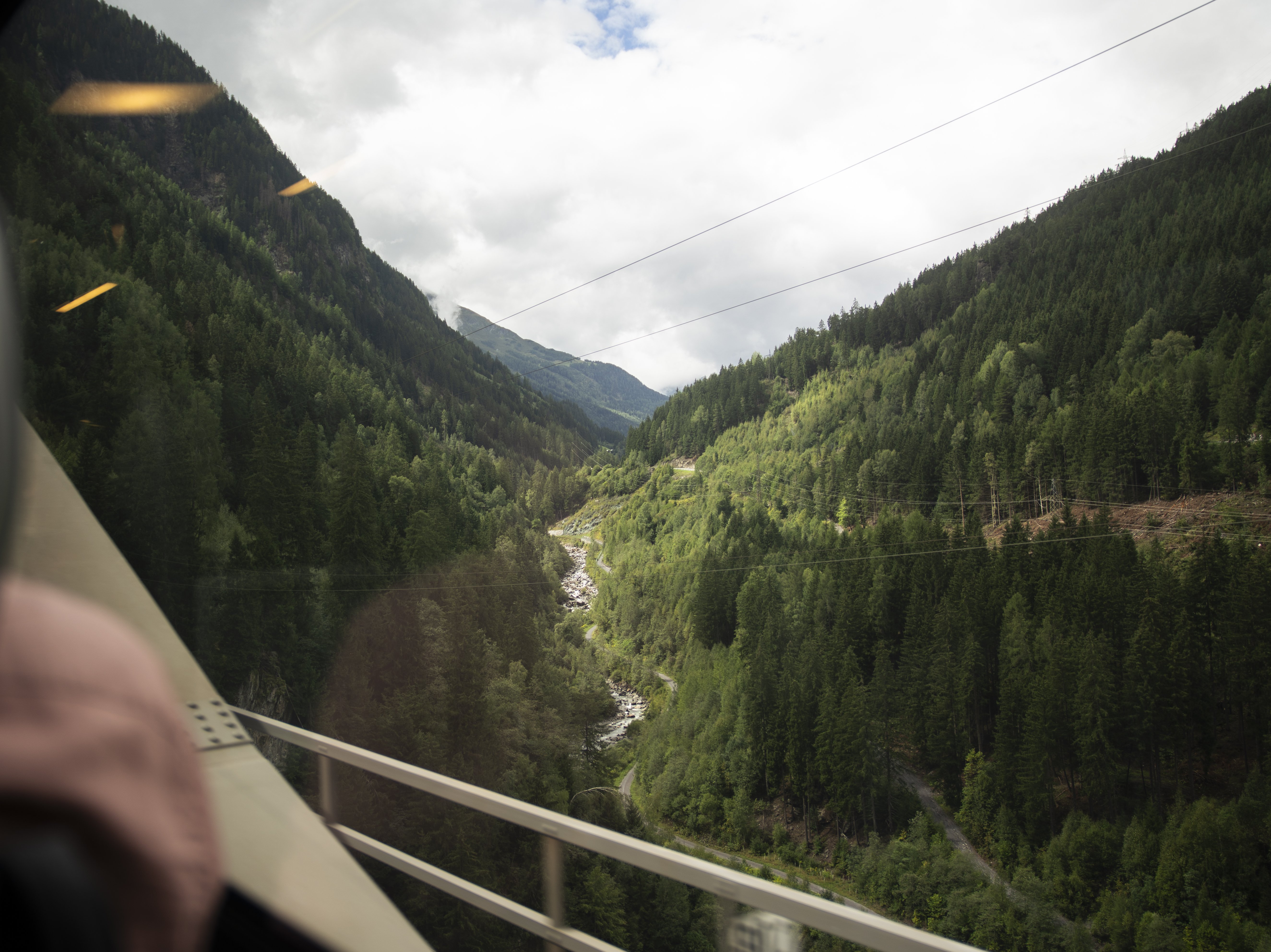 View from a train window of a lush green valley, river below, and forested mountains under a cloudy sky. Part of the train interior is visible on the left side.