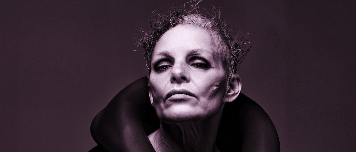 Androgyne Person mit scharfen Wangenknochen, strukturiertem kurzem Haar und einem dramatischen dunklen Outfit mit hohem Kragen starrt selbstbewusst in die Kamera. Das Bild hat einen monochromen violetten Farbton.