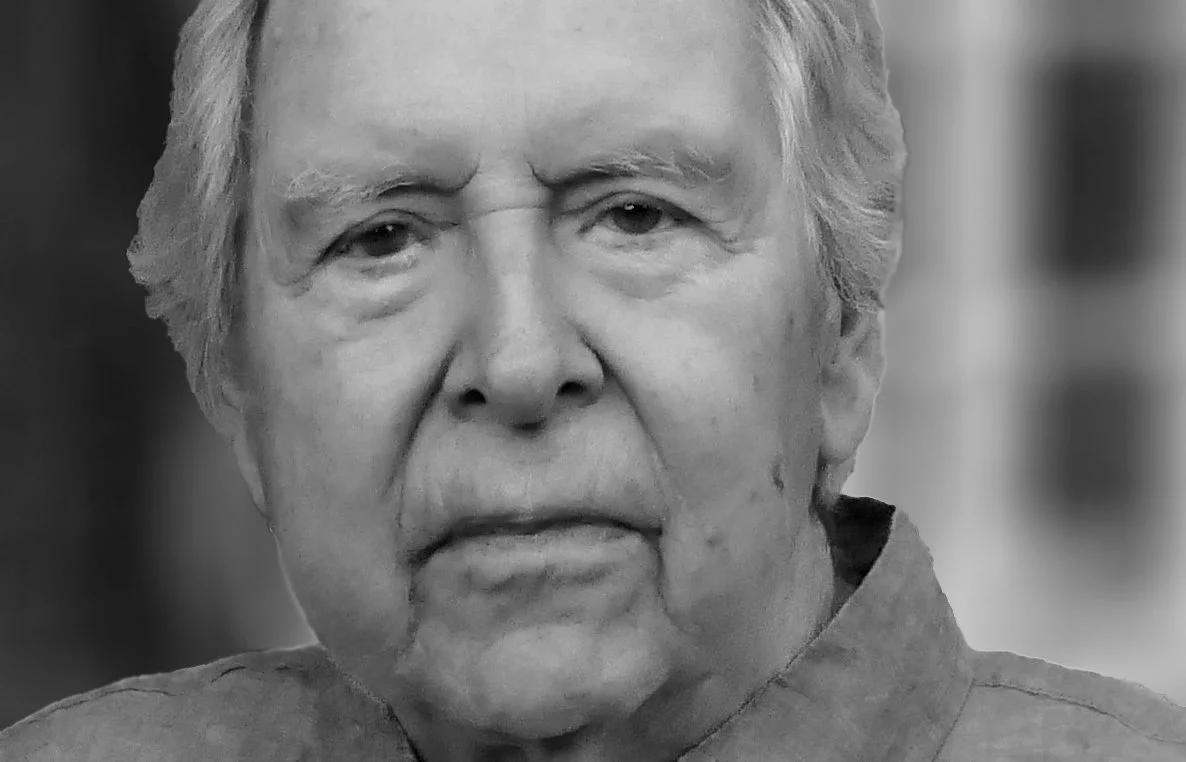 An elderly man with light-colored hair wearing a button-up shirt looks directly at the camera. The background is softly blurred, drawing attention to his face. The image is in black and white.