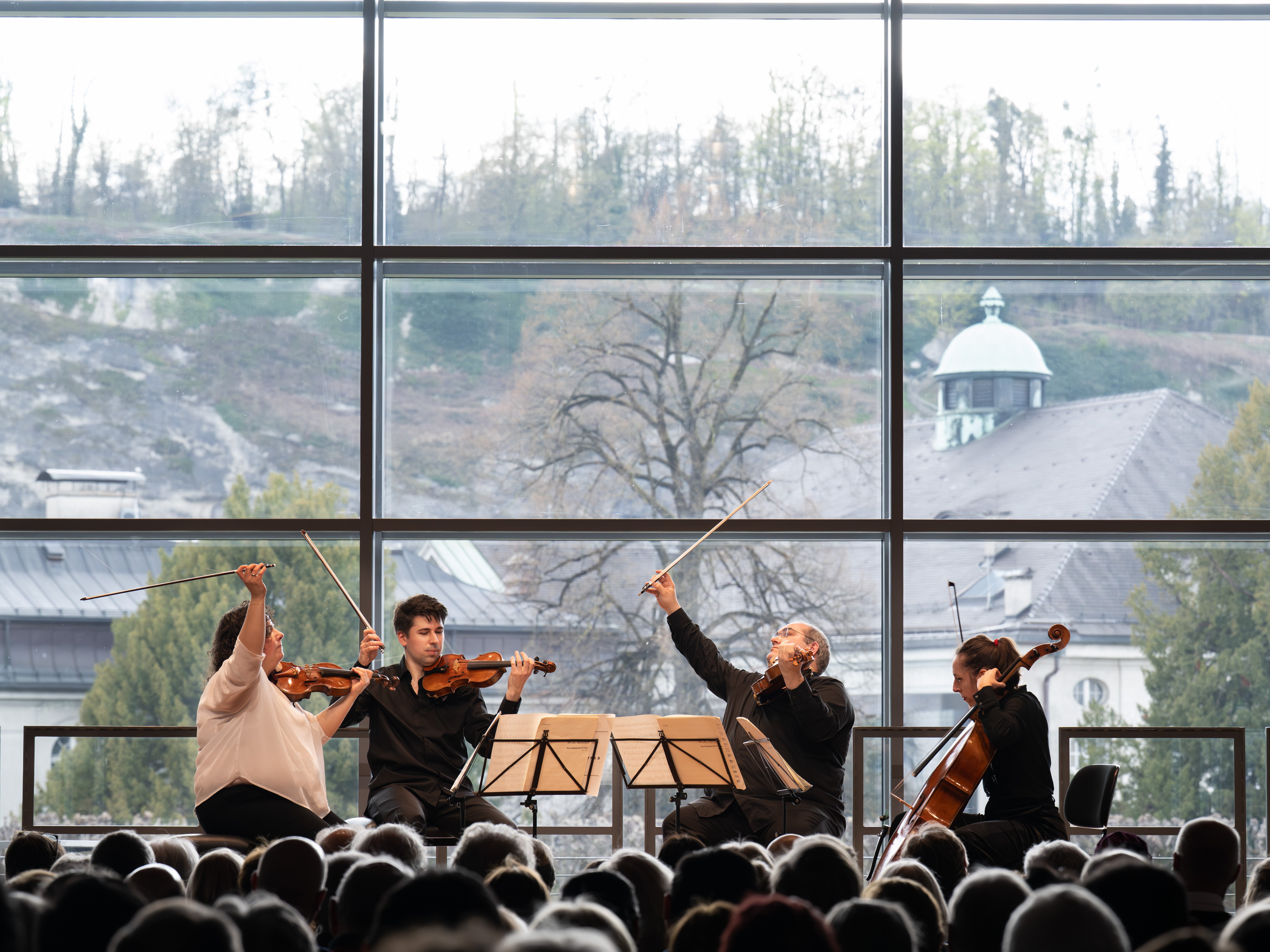 Ein Streichquartett spielt auf der Bühne vor einem großen Fenster mit Blick auf Bäume und Gebäude; ein Publikum beobachtet die Musiker.