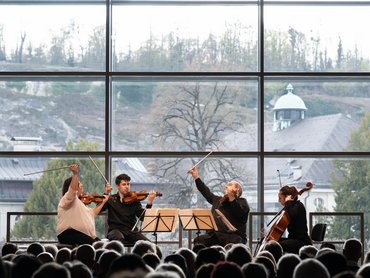 Ein Streichquartett spielt auf der Bühne vor einem großen Fenster mit Blick auf Bäume und Gebäude; ein Publikum beobachtet die Musiker.