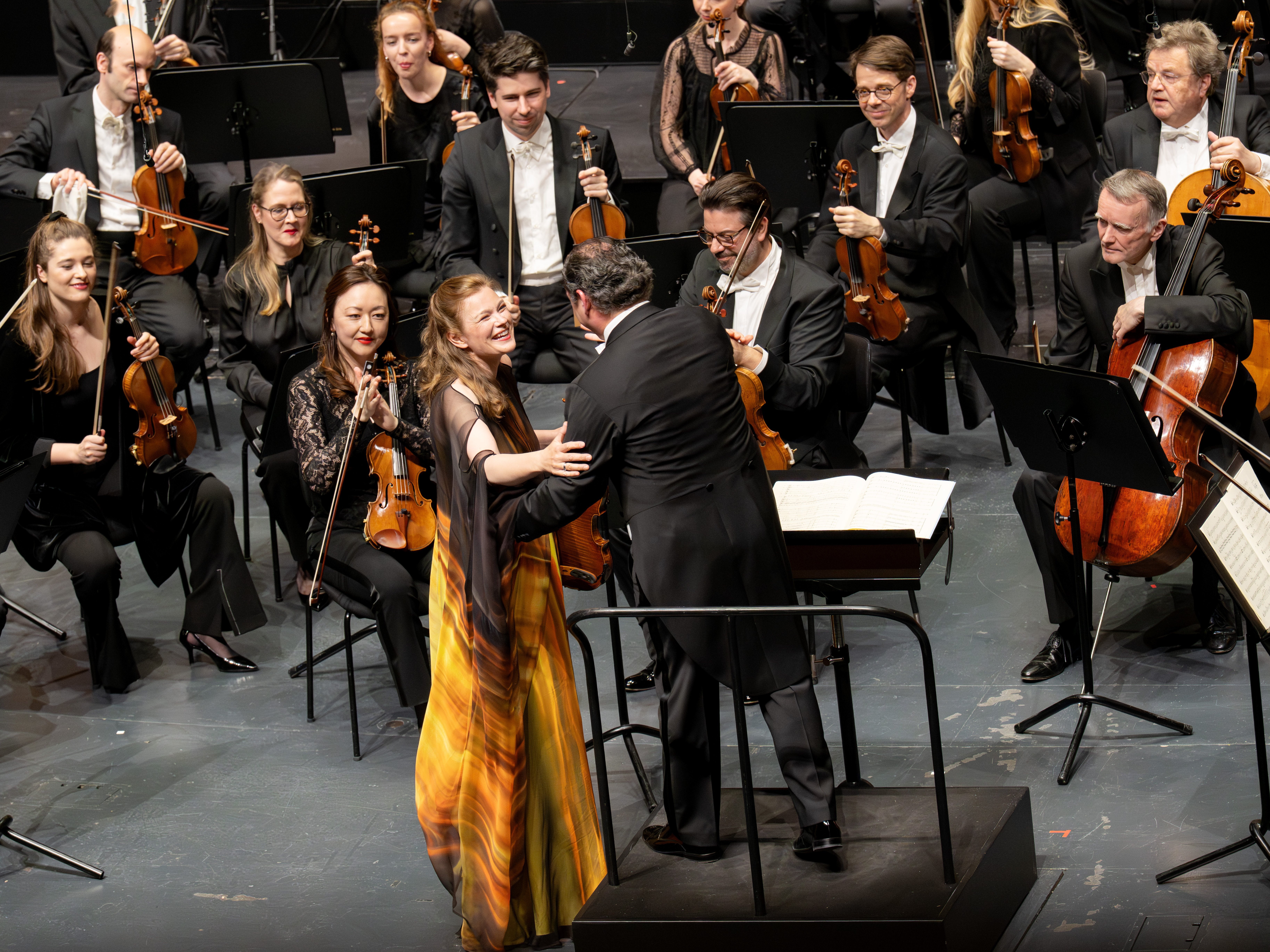 Eine lächelnde Solistin in einem gelben Kleid hält mit einem Dirigenten auf der Bühne Händchen, umgeben von sitzenden Orchestermusikern mit Streichinstrumenten, die alle engagiert und fröhlich wirken.