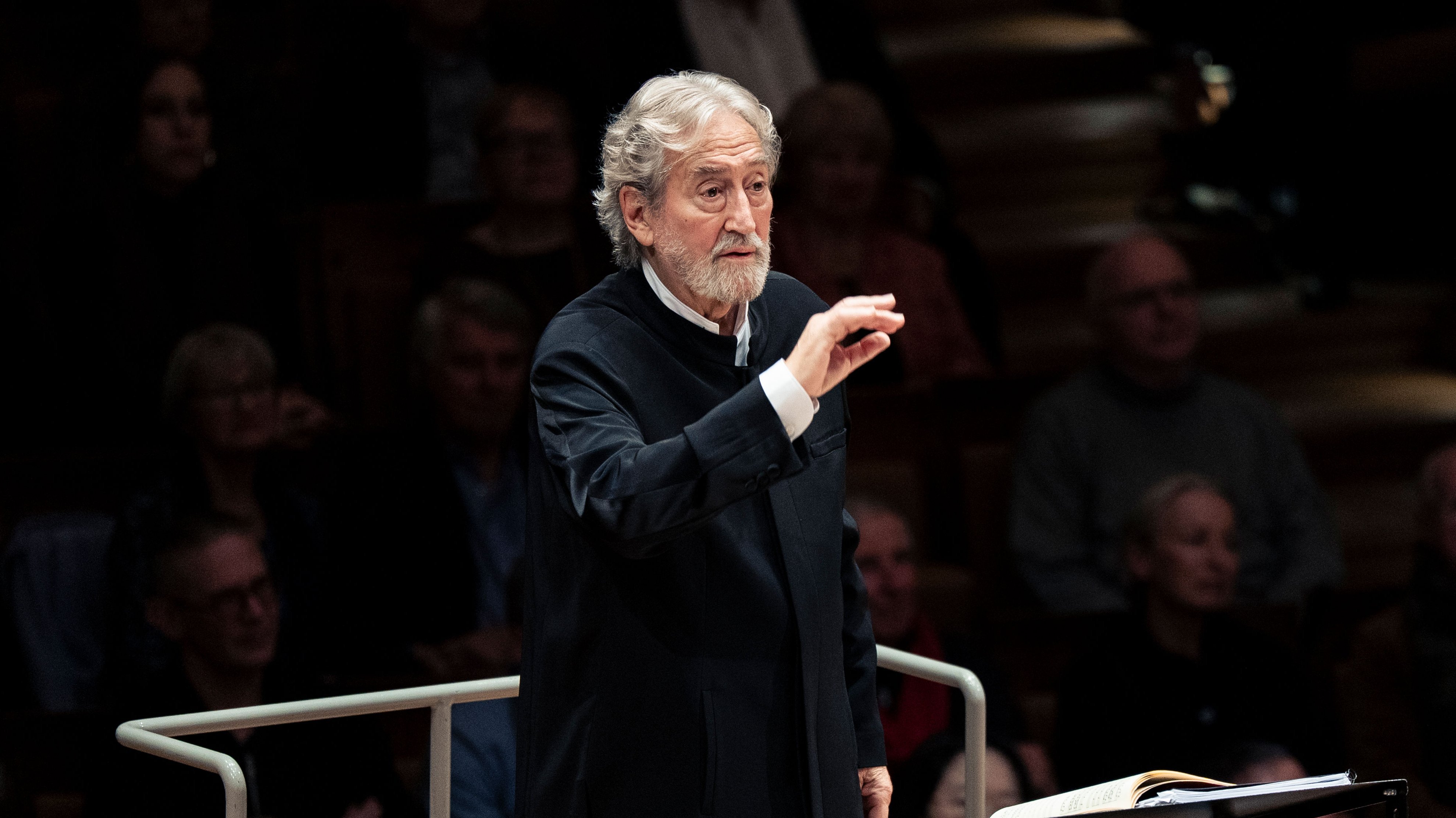 Ein älterer männlicher Dirigent in einem dunklen Anzug leitet ein Orchester auf der Bühne, mit Musikern und Streichinstrumenten im Vordergrund und einem sitzenden Publikum im Hintergrund.
