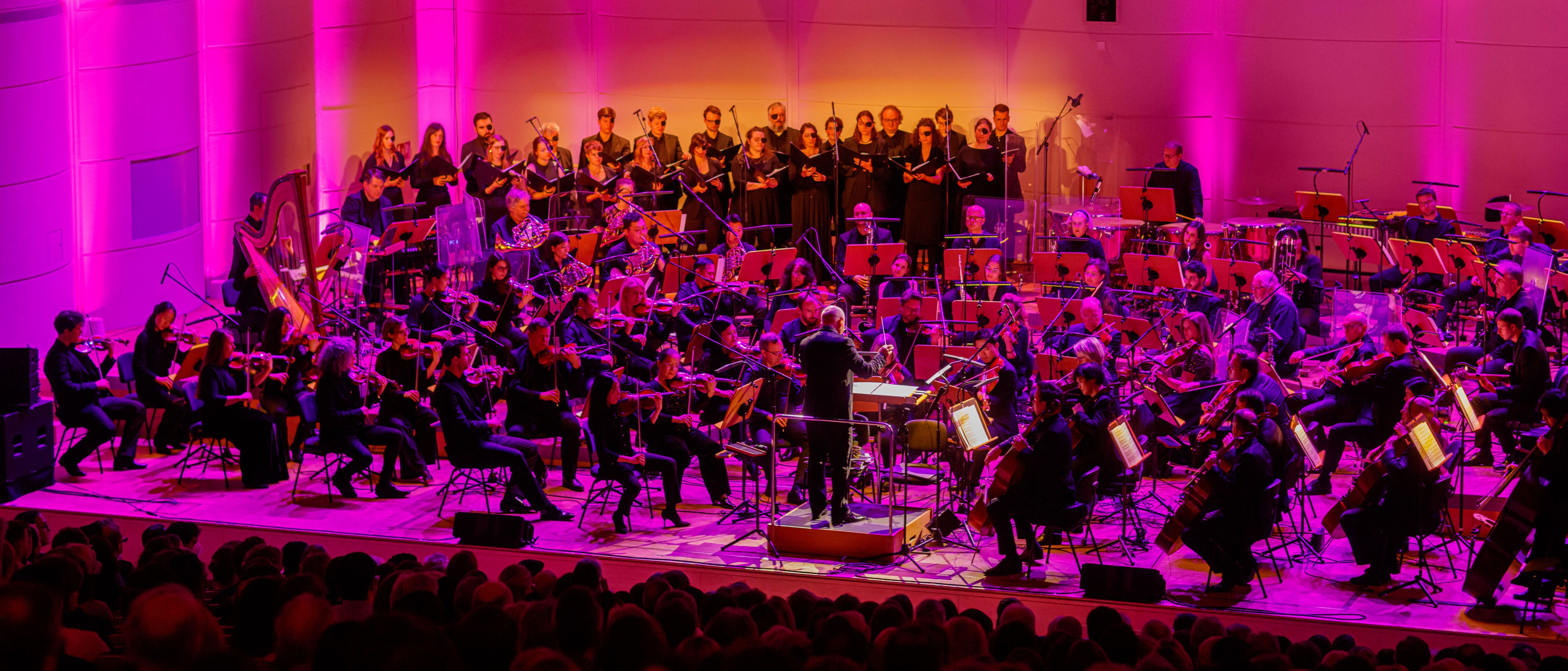 A full orchestra and choir perform on a brightly lit stage with pink and purple lights, in front of a large audience in a concert hall.