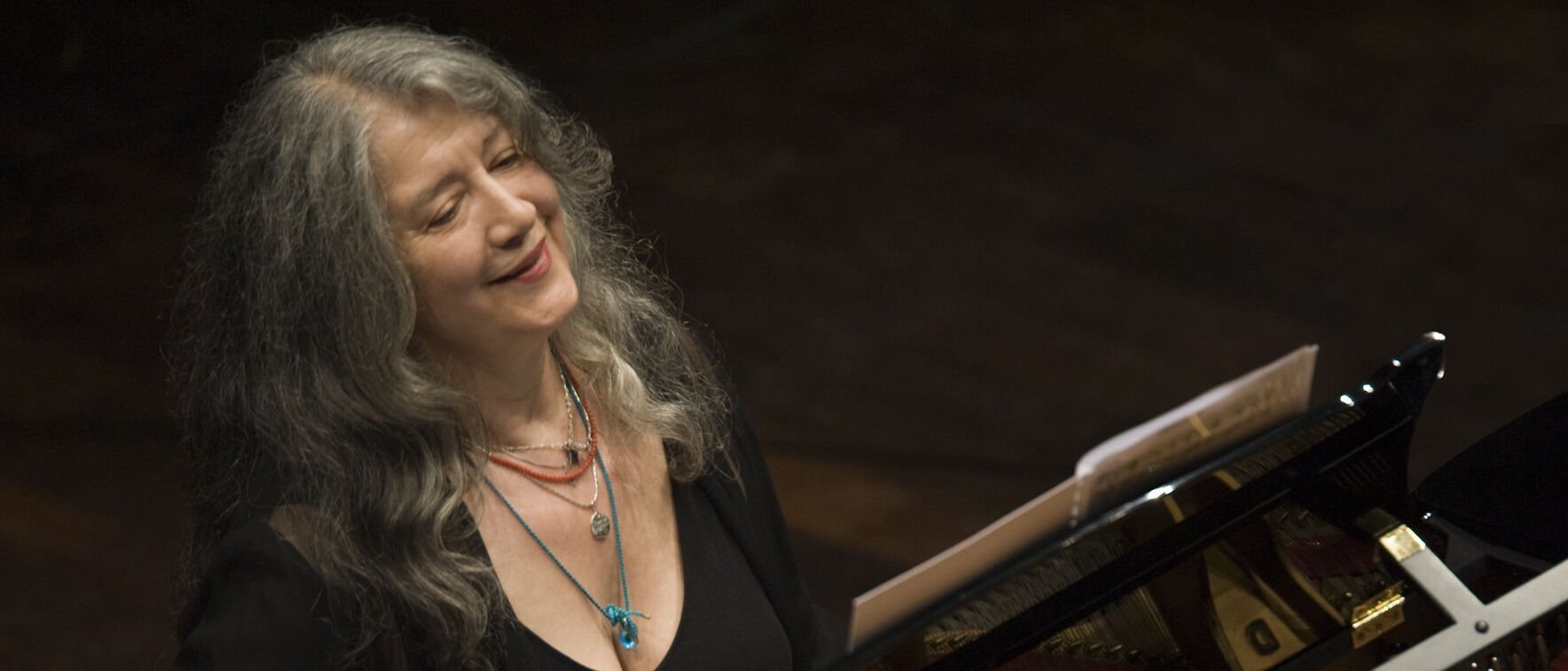 Martha Argerich mit langen grauen Haaren sitzt lächelnd an einem Flügel, trägt mehrlagige Halsketten und ein schwarzes Oberteil. Auf dem Flügel ist ein Notenblatt zu sehen, und die Szene deutet auf eine musikalische Darbietung hin.