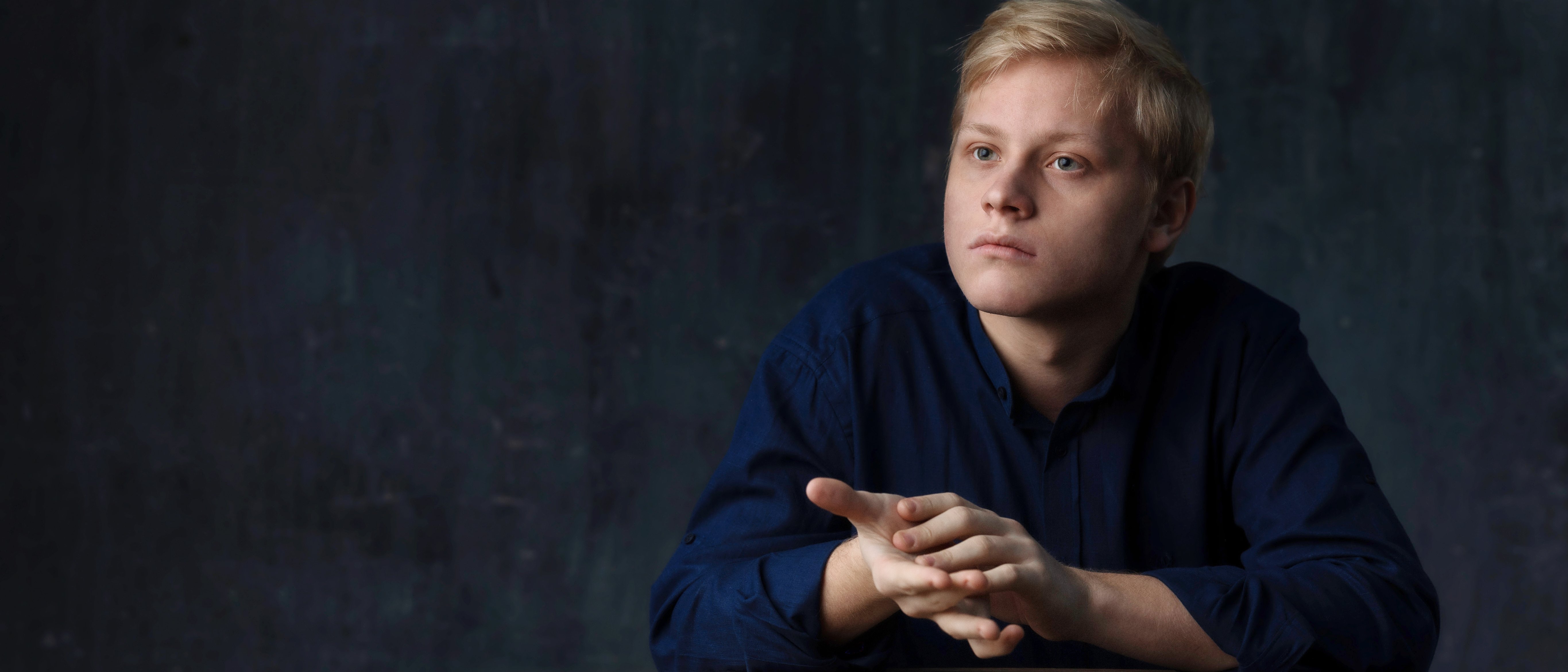 Alexander Malofeev mit blondem Haar und dunkelblauem Hemd sitzt mit gefalteten Händen an einem Holztisch und blickt nachdenklich zur Seite vor einem dunklen, strukturierten Hintergrund.