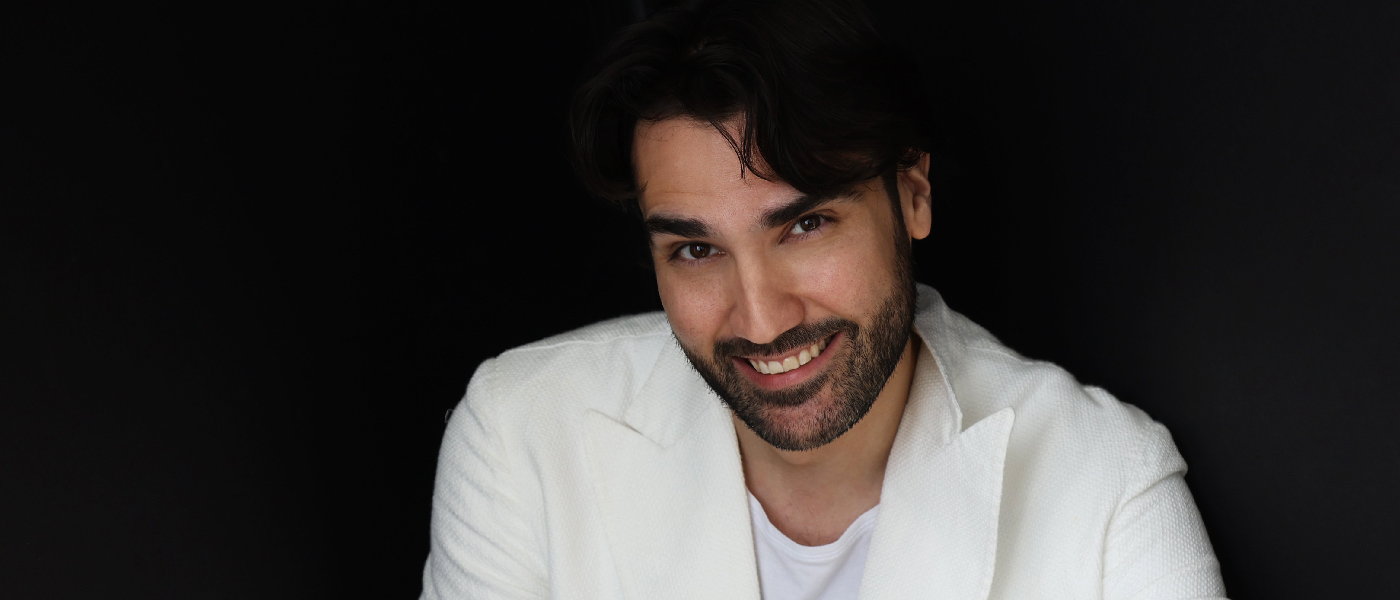 A smiling man with a beard wearing a white double-breasted blazer and white t-shirt against a black background.