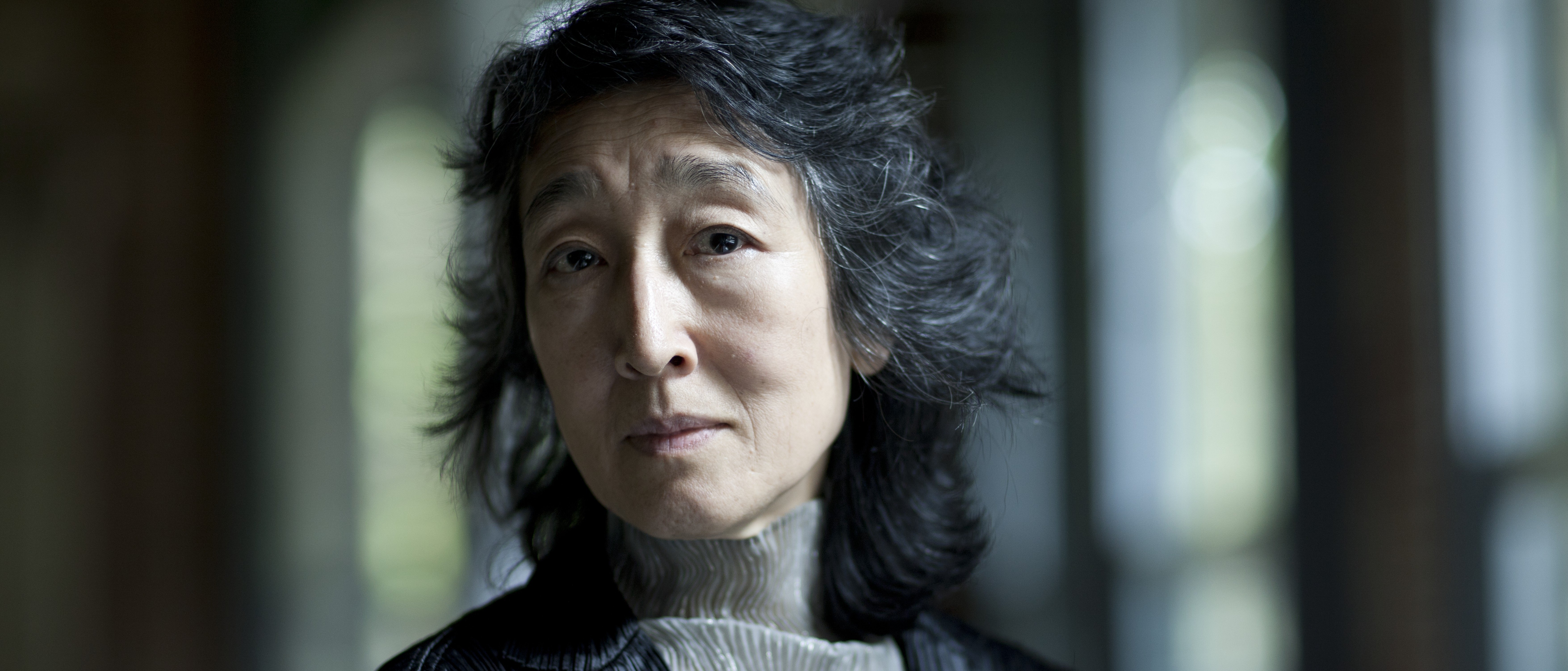 Eine Frau mit gewellten grauen Haaren, die eine schwarze Jacke und eine helle hochgeschlossene Bluse trägt, blickt nachdenklich in die Kamera, während sie in einem Innenraum vor einem leicht verschwommenen Hintergrund steht.